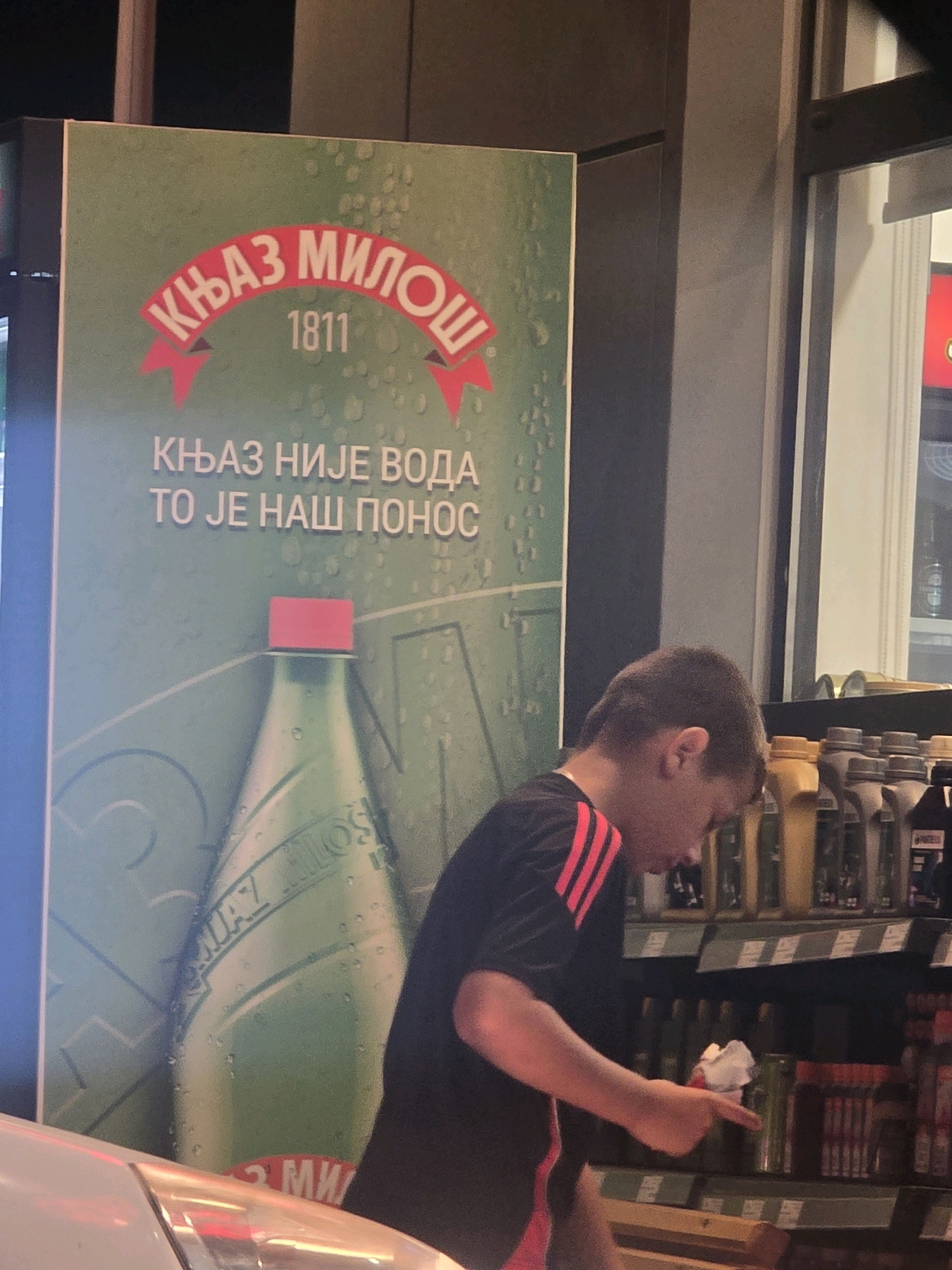 In Podgorica, Montenegro, a young person wearing a black t-shirt with red stripes on the sleeves is reaching for an item on a store shelf. They are standing in front of a large green advertisement poster displaying a bottle of "Knjaz Milos" water and text in Serbian which translates to "Knjaže, you are not water, you are our pride." The poster also shows the year "1811." The store appears to be a retail environment with shelves stocked with various products. The lighting suggests it is either evening or an indoor commercial space.