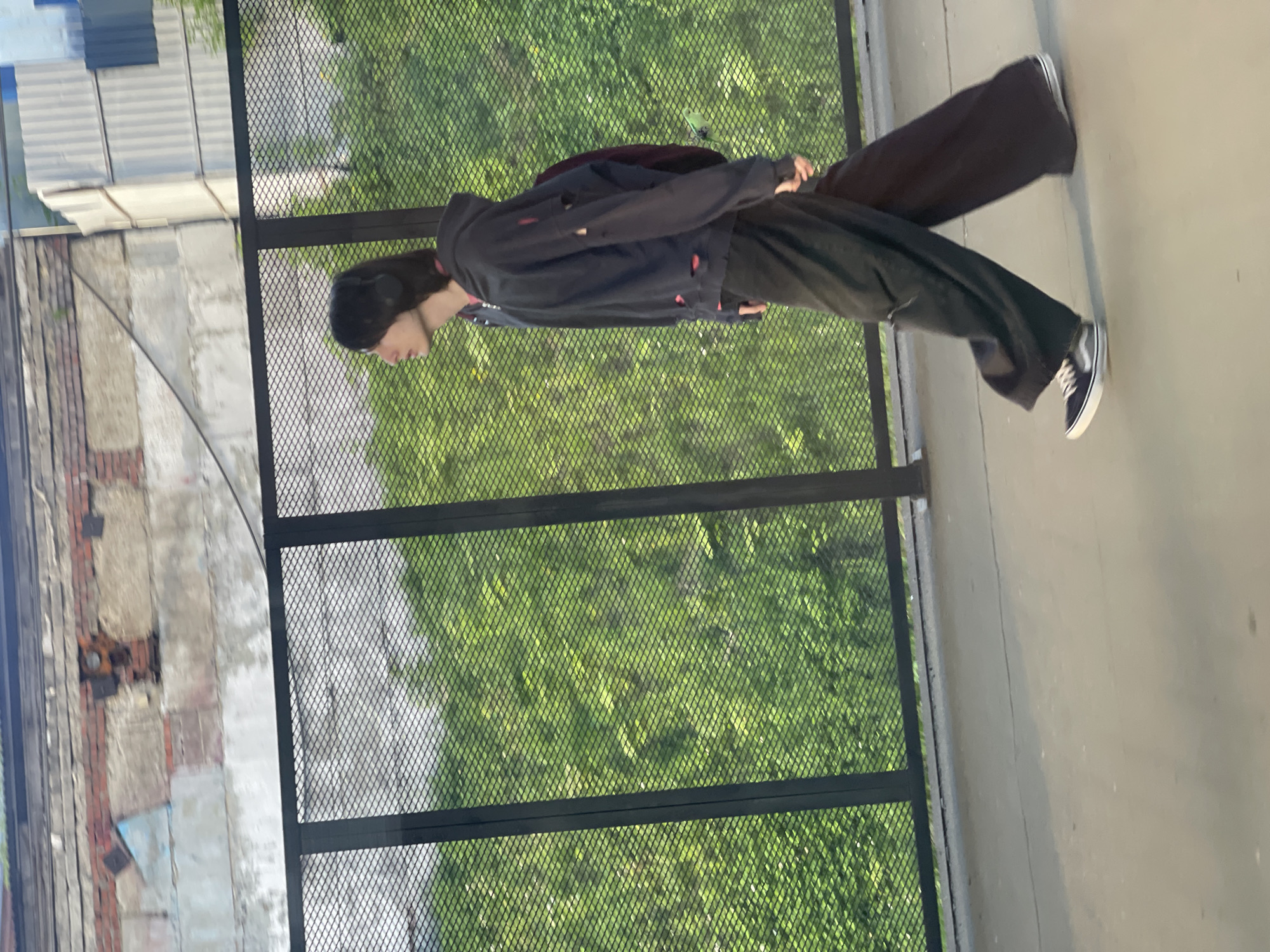 A person is standing outside a metal fence in Khimki, Russia. The person is wearing a black hooded sweatshirt, dark pants, and black and white sneakers. The person is also wearing headphones. Beyond the fence, there is lush green vegetation. To the left of the fence, there is a brick building with visible signs of wear and tear. The setting is outdoors, likely on a paved area next to a building and overlooking a green space. The image appears to be taken during daylight hours.