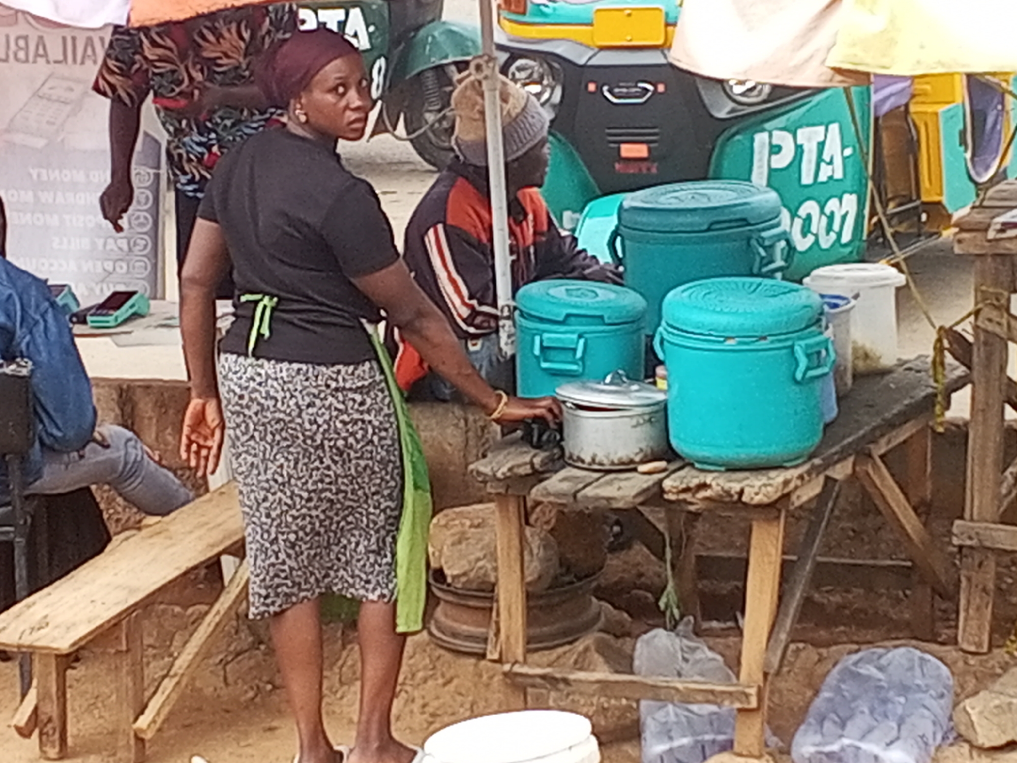 The image depicts a street scene in Gwong, Nigeria. A woman with a maroon headscarf, wearing a black shirt and a patterned black and white skirt with a green apron, stands near a makeshift wooden table. She is looking over her shoulder. On the table are several turquoise containers, a metal pot, and a white plastic bucket. A man wearing a gray beanie and a jacket with orange and white stripes sits behind the table.
In the background, a three-wheeled vehicle, known locally as a "Keke Napep," is partially visible. The vehicle has "PTA" written on it. Another man wearing a patterned top is seen to the left of the woman. A wooden bench is visible to the left, where another person is seated.