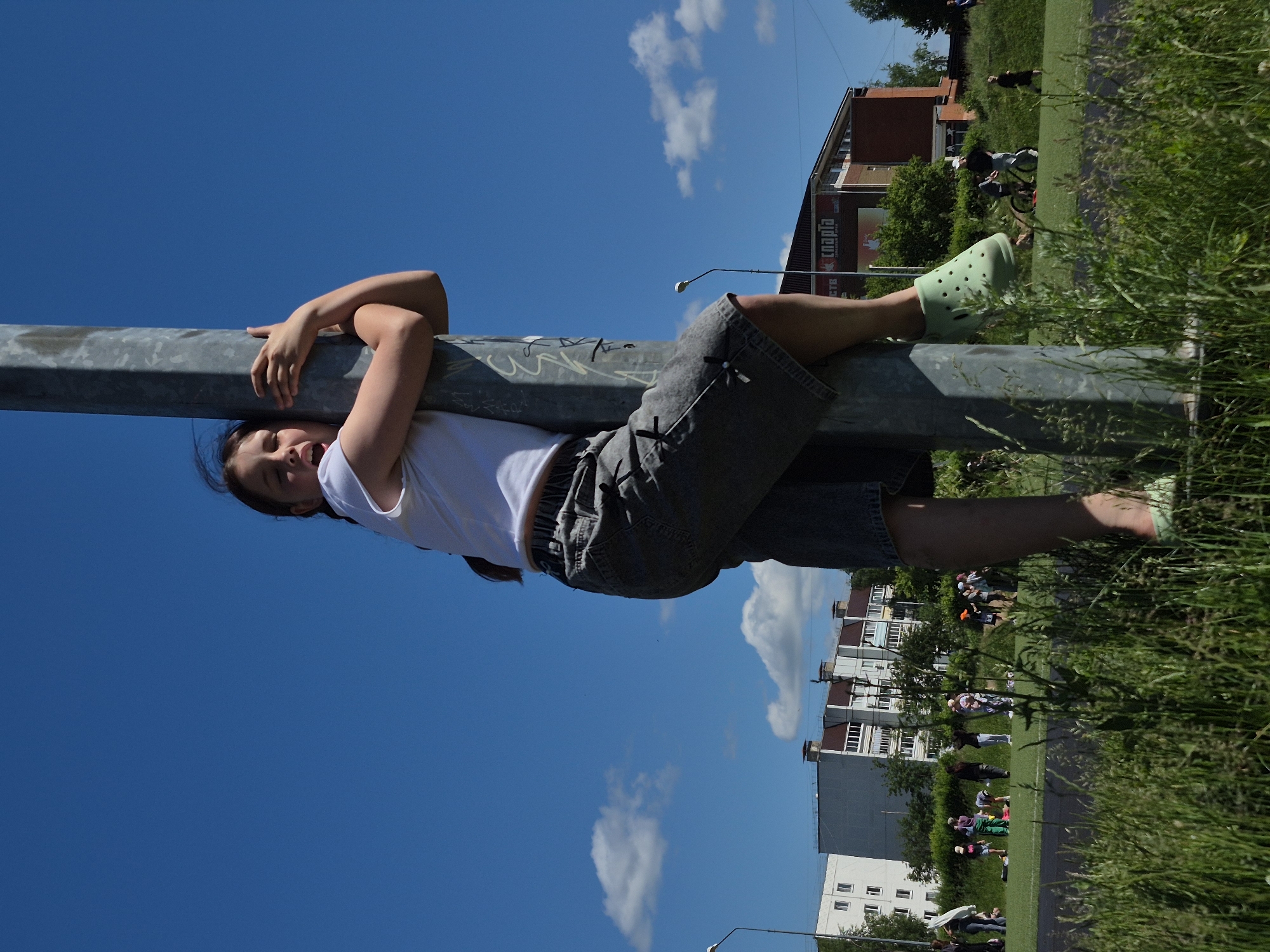The media file shows a young person hanging upside down from a metal pole outdoors on a sunny day. The subject is wearing a white t-shirt and grey shorts. Their arms are wrapped around the pole, and their legs are extended upwards. They are looking towards the camera with a playful expression. Light green Croc-style shoes are visible on their feet.
The background consists of a bright blue sky with scattered white clouds. In the distance, there are apartment buildings and trees, suggesting an urban or suburban setting. A strip of green grass is visible in the foreground, with some of it obscuring the lower part of the frame. The lighting indicates it is daytime, likely afternoon.
The overall scene suggests a moment of recreation or playful activity. The image is oriented sideways. Some graffiti is visible on the pole. A sign with Russian text, possibly advertising "Chaikovskaya," is partially visible on one of the buildings.