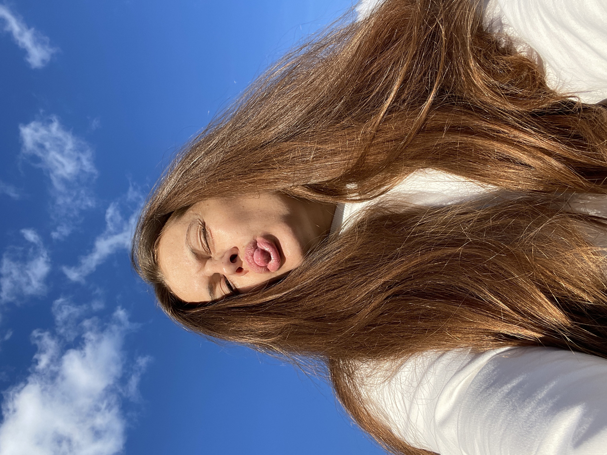 A young woman with long, brown hair is pictured outdoors against a bright blue sky with scattered white clouds. She is looking upwards, with her eyes closed and her tongue sticking out playfully. Her face is partially obscured by her hair, which frames her face and extends across the frame. She is wearing a white top. The lighting suggests it is daytime, likely a sunny day. The image appears to be a selfie, taken from a low angle looking up at the sky. The provided location context is Zarechny, Russia. There is no visible text in the image.