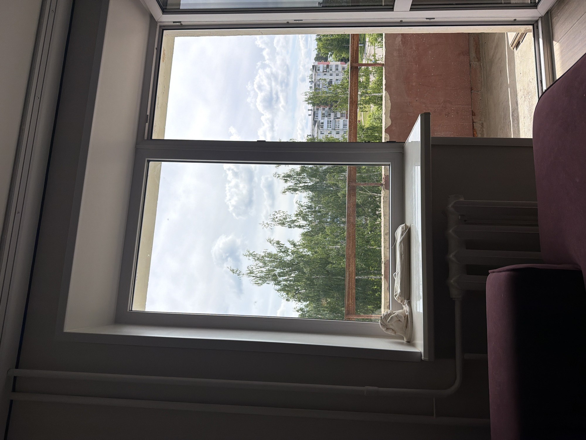 The image captures an interior view looking out through a window. The overall scene, particularly the exterior elements, appears rotated 90 degrees counter-clockwise within the frame, suggesting the photograph was taken with the camera held sideways.
The window features white frames, dividing it into two main panes, one above the other. Through the window, an outdoor scene is visible. Directly beyond the window is a balcony with a rusted metal railing. The balcony floor is concrete, and its wall is a reddish-brown color, possibly brick or painted concrete. Beyond the balcony, a vibrant line of green trees dominates the mid-ground. In the distance, several multi-story white residential buildings with multiple windows are visible, characteristic of Eastern European architecture, consistent with the given location context of Chaikovsky, Russia. The sky above is bright and filled with large, fluffy white cumulus clouds.
Inside the room, the walls are a light, likely white or off-white, color. A white window sill or ledge runs along the bottom edge of the lower window pane. Draped over the window frame and resting on the sill is a white, fabric-like object that resembles a pair of cupped hands. Below the sill, a white, ribbed radiator is affixed to the wall, connected by white pipes. On the far right side of the image, a portion of dark red or burgundy upholstered furniture, likely an armrest of a sofa or armchair, is visible.
The scene depicts a calm daytime view, with bright, cloudy weather. No specific activity is occurring, and there is no visible text.