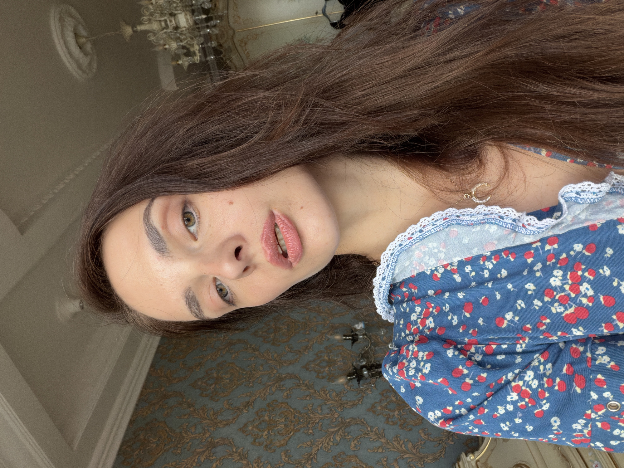 A woman with long, dark brown hair is shown from the chest up, taking a selfie. She is wearing a blue dress with a red and white floral pattern and a white lace trim. A gold necklace with a crescent moon pendant is visible. The background includes a wall with ornate patterned wallpaper and a chandelier hanging from the ceiling. The lighting suggests an indoor setting, possibly a room in a house or hotel in Ufa, Russia.
