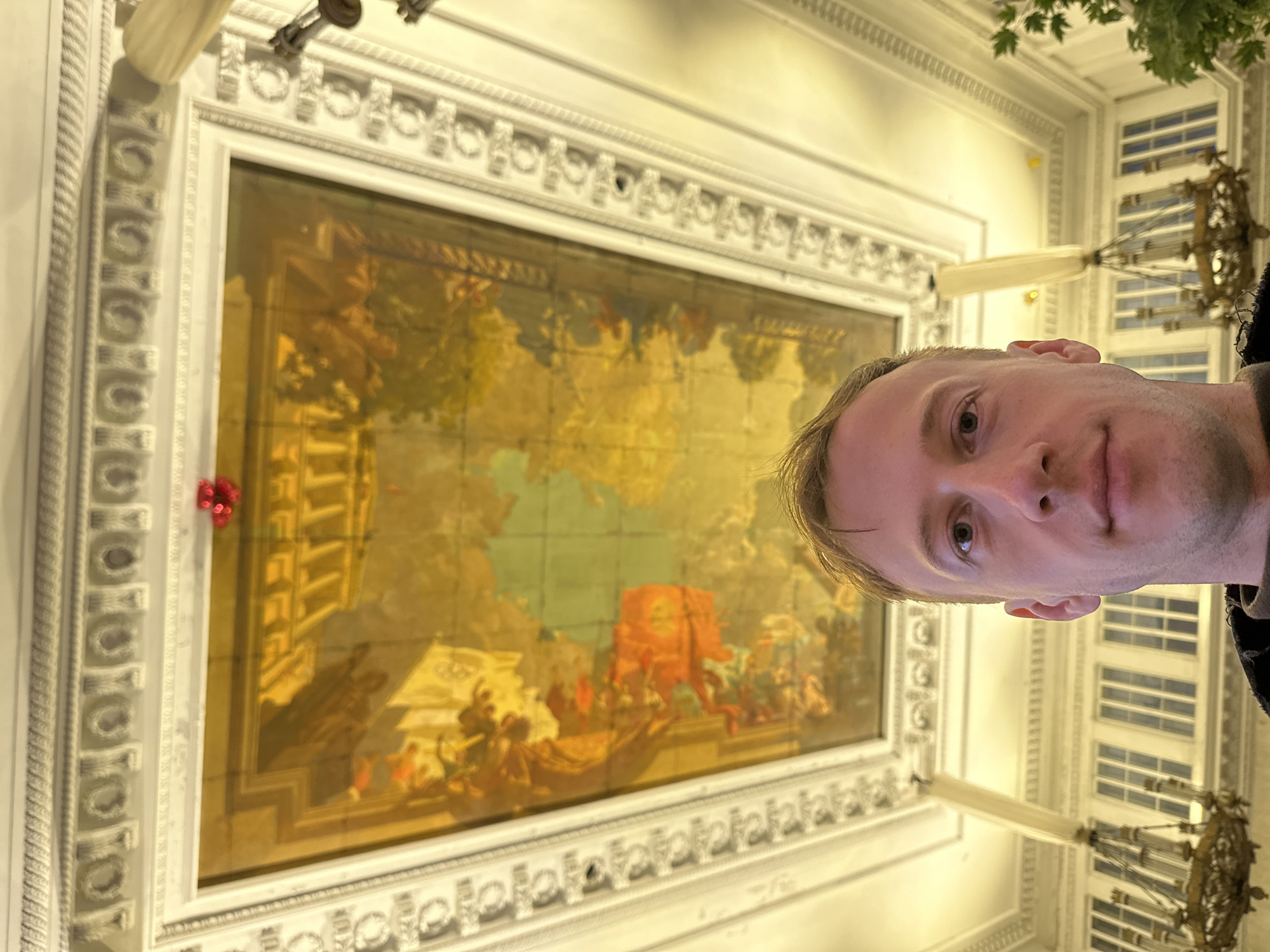 The image depicts a person with light skin and short blond hair, facing the camera at an angle, seemingly taking a selfie. The background reveals the ceiling of a grand building in Saint Petersburg, Russia, specifically the ceiling artwork of the State Hermitage Museum, which is visible behind the person. The ceiling art is framed with ornate, white molding. Chandeliers are visible hanging from the ceiling.