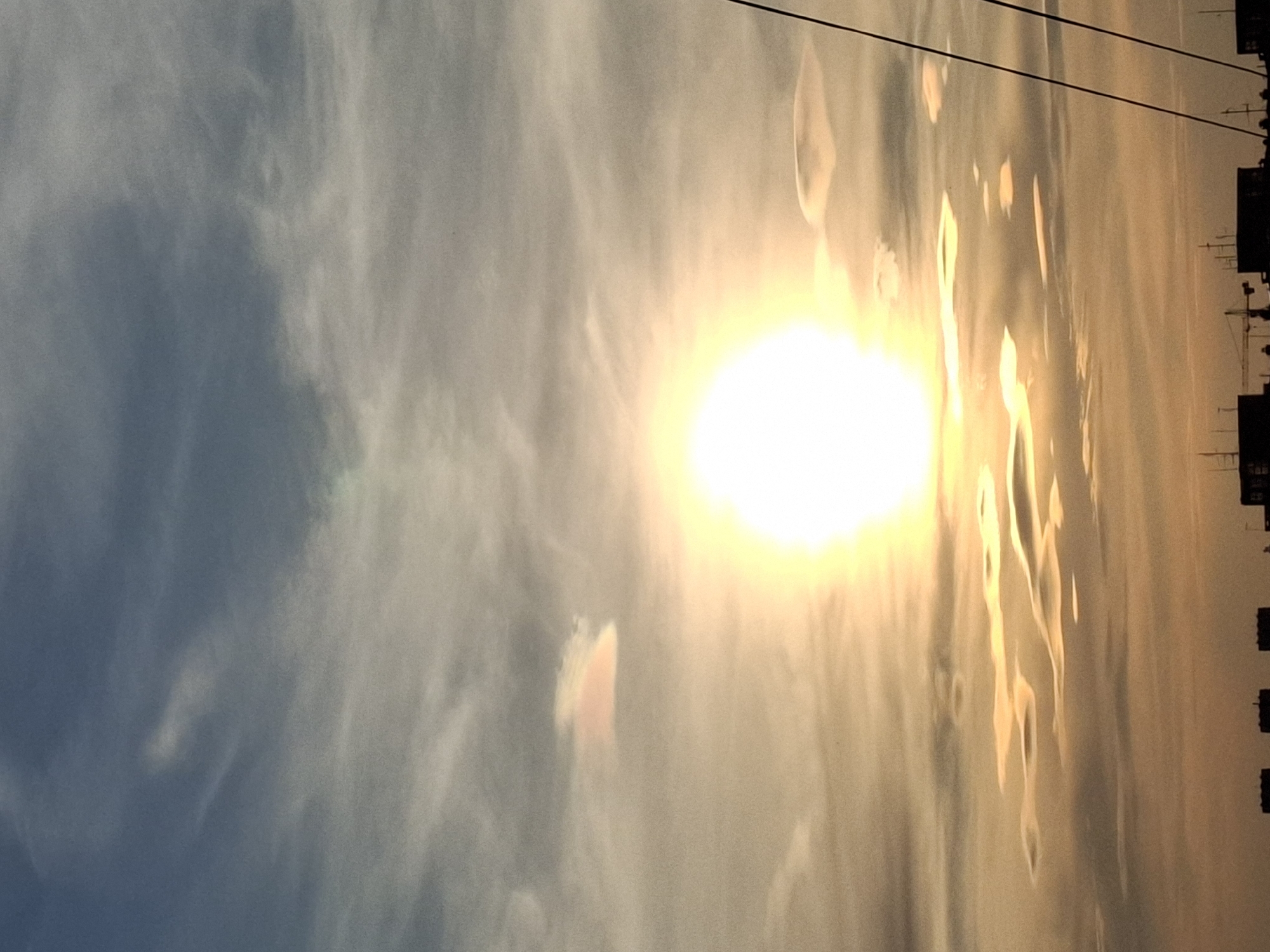 The image depicts a bright sun in a hazy sky, likely at sunset or sunrise. The sky transitions from a blueish hue on the left to a yellowish-orange near the sun. The edges of buildings are silhouetted against the sky, and utility lines run across the upper portion of the frame. There are reflections or light patterns creating vertical lines through the sky and possibly water in the sky. The image's location is likely Novi Sad, Serbia.