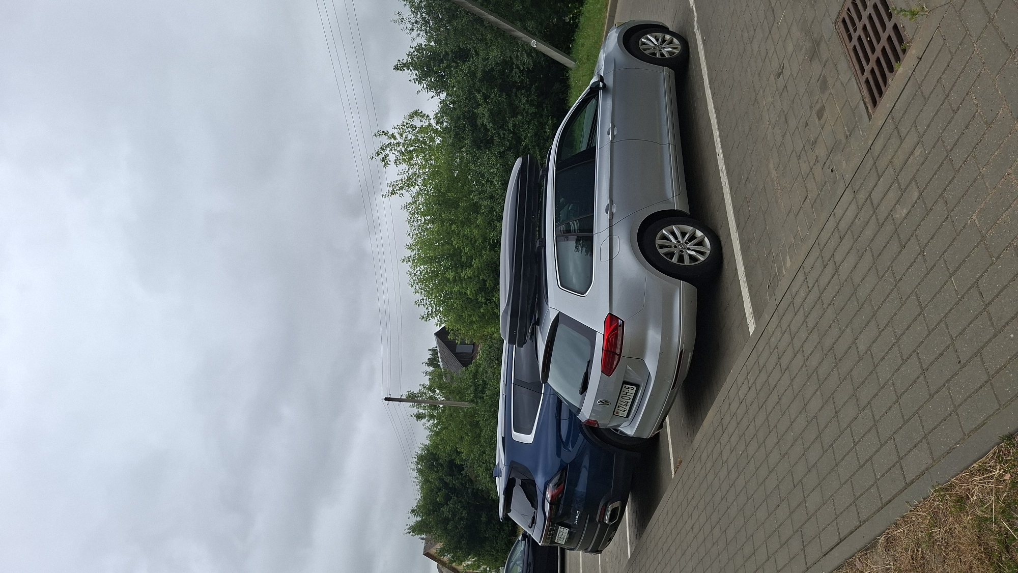 Two parked cars are visible in a paved parking lot. The car in the foreground is a silver Volkswagen Passat Variant, equipped with a black cargo box on its roof. The license plate of this car reads "BY 4744 OH-5". To its left, a dark blue SUV, possibly a Geely Monjaro, is parked. The SUV's license plate is partially visible and reads "BY 9955 EM-5". In the background, a line of lush green trees separates the parking lot from houses and utility poles, indicating a suburban or rural setting. The sky is overcast and appears to be cloudy.