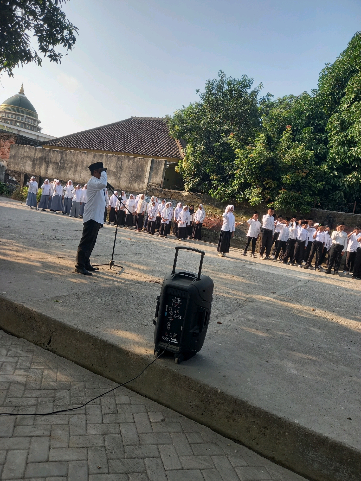 The image depicts an outdoor scene with a group of students, predominantly female, dressed in white shirts and hijabs, lined up and seemingly participating in a ceremony or assembly. A male figure in a white shirt and black pants, wearing a black peci (cap), stands in front of a microphone on a stand, appearing to address the students. A large black speaker is positioned on the ground to the right of the frame. The background includes a building with a domed roof, suggestive of a mosque, and lush green trees. The ground is paved with concrete and interlocking bricks. The lighting indicates it is daytime, likely morning or late afternoon, with shadows cast by the subjects. The overall setting appears to be a school or religious institution in Perampuan, Indonesia.