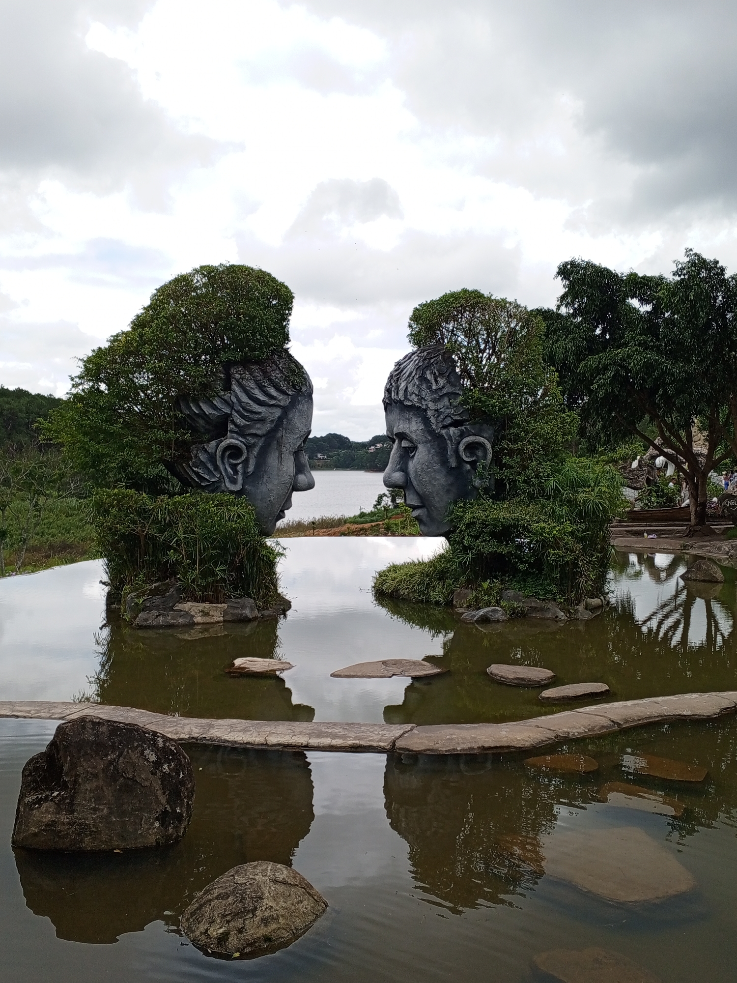 The image shows a surreal landscape sculpture in Da Lat, Vietnam, featuring two colossal stone heads integrated with lush greenery, facing each other across a calm body of water. The sculptures, designed to resemble human faces, are partially covered in dense foliage, with trees growing from the tops of their heads and smaller shrubs adorning their sides. These structures are situated in a man-made pond or reflection pool, with flat stones forming a pathway that appears to float on the water's surface. The background reveals a serene natural setting with rolling hills, distant trees, and a cloudy sky that casts a diffused light over the scene. The overall impression is one of art blending seamlessly with nature, creating a tranquil and thought-provoking atmosphere.