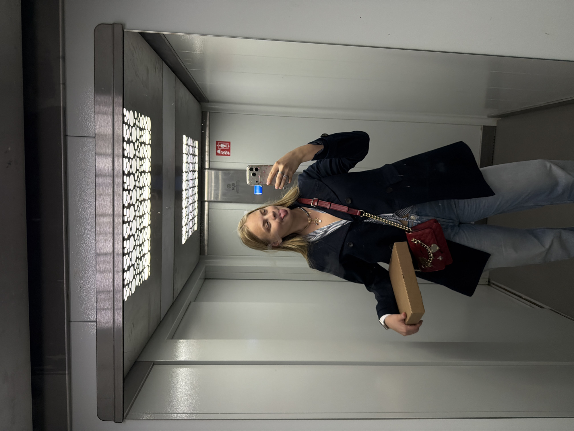 A young woman with long blonde hair is captured in a mirror selfie inside an elevator in Izhevsk, Russia. She is dressed in a dark blazer over a striped shirt and light-colored jeans. Over her right shoulder, she carries a red quilted shoulder bag with a gold chain strap. In her left hand, she holds a plain brown cardboard box. She is taking the picture with a light-colored smartphone, held up to the mirror. Her expression is playful, with her tongue slightly extended.
The elevator interior appears modern, featuring metallic panels on the walls. A distinctive patterned light panel, composed of illuminated white circles, is visible on the left wall. On the back wall, an emergency panel is present, displaying a red symbol of a person with an arm raised, often indicating an emergency call or alarm button. Below this symbol, some inverted text is visible in the mirror, including what appears to be "M.2.3". The lighting within the elevator is bright and even. The time of day and weather are not discernible.