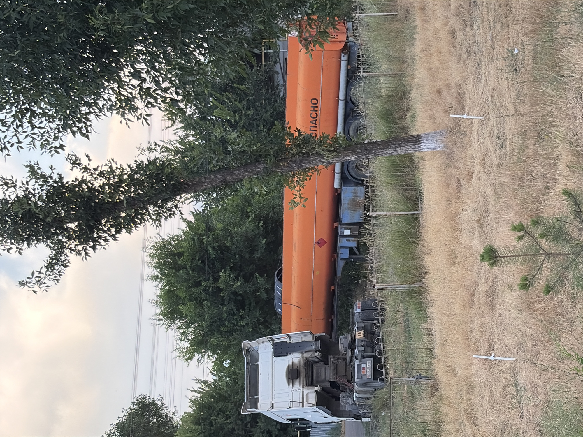 This media file depicts a white truck with an orange tanker trailer parked in a grassy, rural setting. The tanker is labeled with the Cyrillic text "СПАСНО," which translates to "SAVED" or "RESCUE." The truck appears to be a commercial vehicle, possibly a fuel or chemical transporter, given the nature of the tanker. The scene is set outdoors, with trees and dry grass surrounding the vehicles. A wire fence runs along the right side of the frame, and power lines are visible in the distance against a cloudy sky, suggesting it is daytime. The overall impression is one of a functional, if somewhat isolated, location. There are no people visible in the image. The grass appears dry and somewhat unkempt, hinting at either a dry season or a less maintained area. The image is oriented vertically, with the truck and tanker appearing to be on a slight incline or the camera tilted.