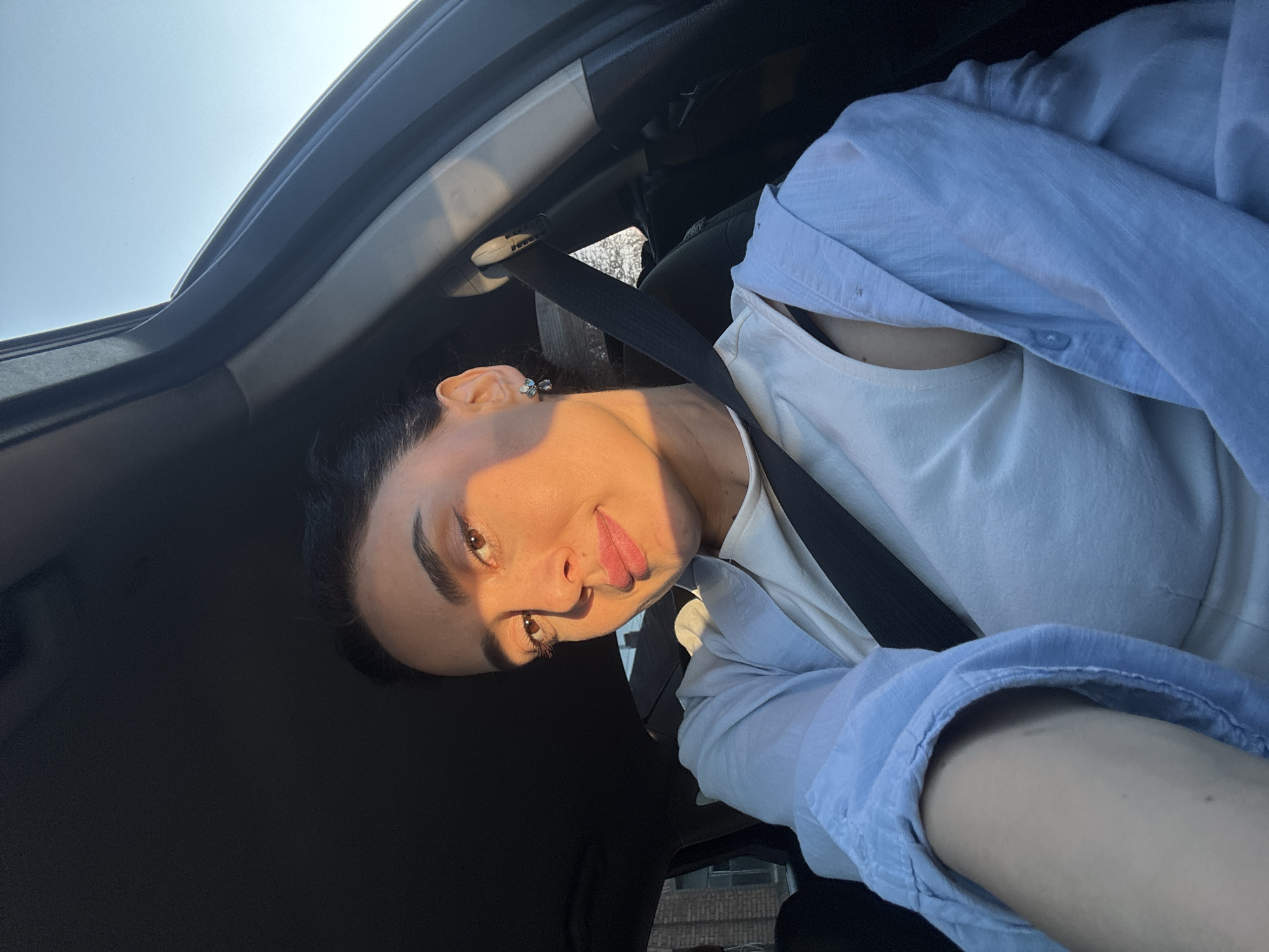 A young woman is captured in a tilted, close-up selfie taken from inside a car, likely in the front passenger seat. She looks directly towards the camera with a subtle, pleasant smile and a confident gaze.
The woman has dark hair, styled away from her face, and prominent eyebrows. Her brown eyes are clearly visible, and she wears a light pink lipstick. A small, metallic earring, possibly featuring a floral or geometric design, adorns her visible ear. She is dressed in a light blue, long-sleeved button-up shirt, worn open over a white undershirt or top. A black seatbelt is fastened across her chest and shoulder.
The setting is the interior of a car, with parts of the dark-colored roof lining, door frame, and the top of a seat visible. The background outside the car window on the left reveals a bright, clear blue sky, while a blurred glimpse of urban architecture can be seen through the lower window on the right. The scene is bathed in a warm, golden light that illuminates the right side of the woman's face and shoulder, strongly suggesting the photograph was taken during the late afternoon or early evening, often referred to as the golden hour. The weather appears sunny and clear.
The image conveys a relaxed and casual mood. No visible text is present in the media. The location context provided is Tbilisi, Georgia.