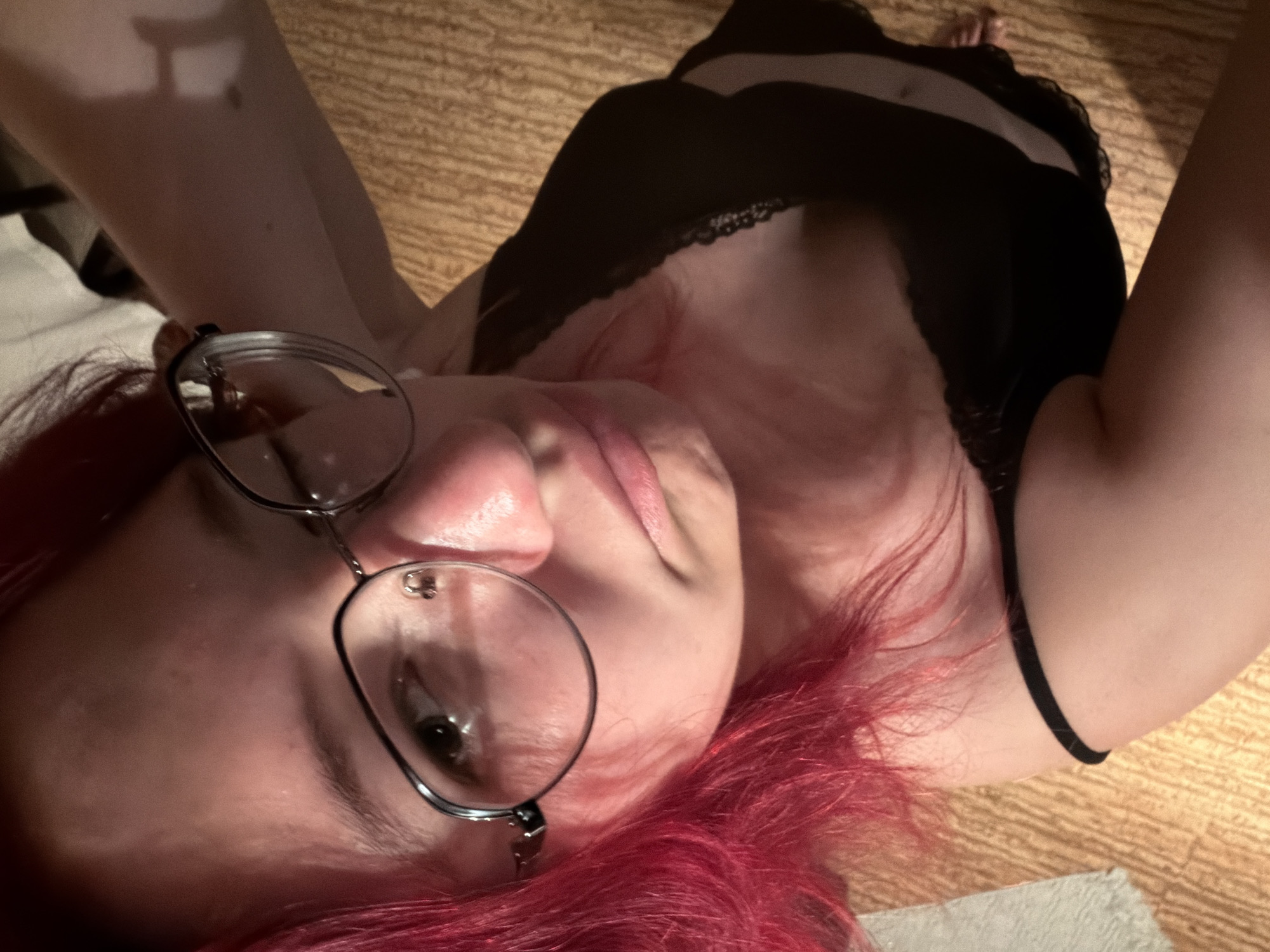 The image is a close-up, high-angle selfie of a young person with bright pink hair. They are wearing a black camisole with lace trim and glasses with thin wire frames. The person's face is partially obscured by their hair and glasses, and they appear to be looking down with a slightly pursed expression. The background consists of a textured, light brown surface, possibly a floor or rug, and a hint of a white rug or blanket in the upper left corner. There are no discernible landmarks or elements that would indicate a specific location within Chelyabinsk, Russia. The lighting is dim, casting shadows that emphasize the contours of the person's face and shoulders. The overall impression is of a casual, private moment captured by the individual themselves.