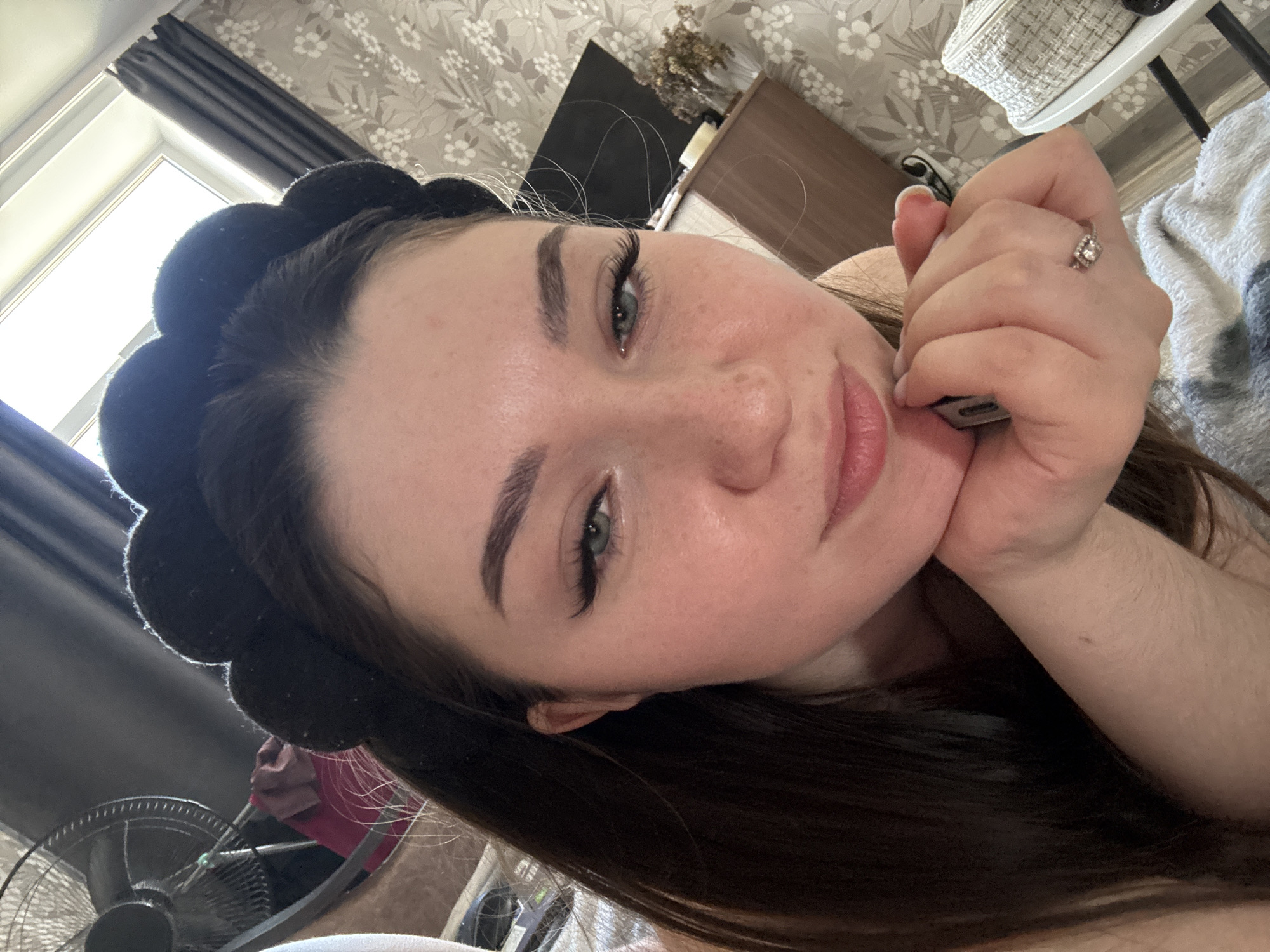 A young woman with long brown hair, wearing a black fuzzy headband, is taking a selfie in what appears to be a bedroom. She is leaning her chin on her hand, which is resting on her chest. She has on makeup, including eyeliner and mascara, and a ring on her ring finger. In the background, there is a fan, a window with curtains, patterned wallpaper, a dresser with a vase of dried flowers, and a wicker bag on a chair. The lighting suggests it is daytime. The location is Zarechnaya, Russia.