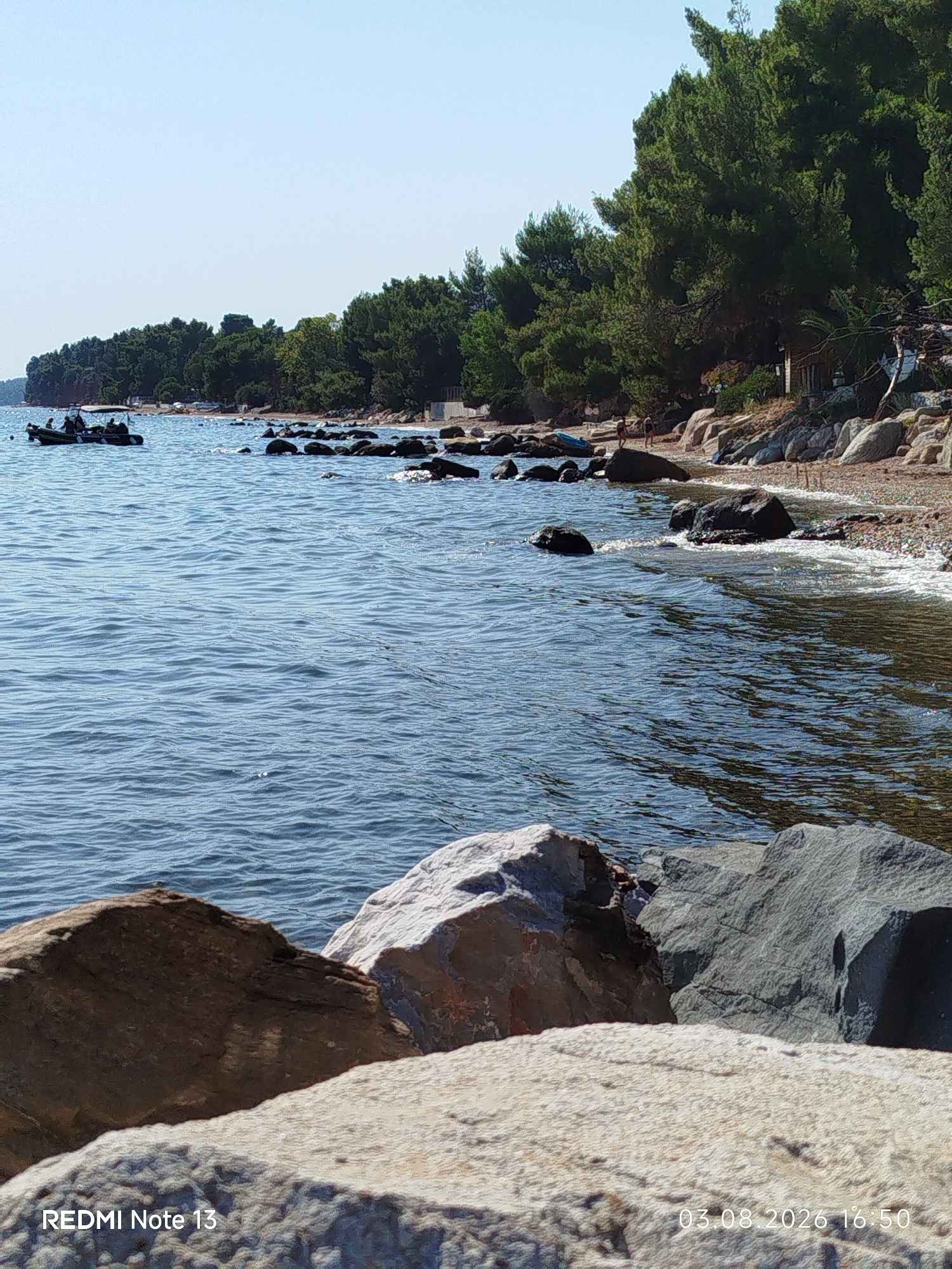 The image depicts a serene coastal scene in Sithonia Municipal Unit, Greece. In the foreground, large rocks are visible, with the sea gently lapping at the shore. In the middle ground, the clear blue water is dotted with several dark rocks. Further back, a tree-lined shore stretches along the coastline, with a small boat carrying people visible on the water. The sky is a clear, bright blue. The overall impression is one of a peaceful summer day at the beach.