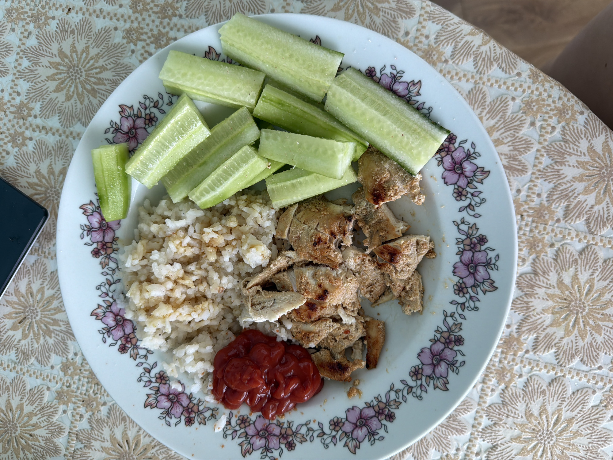 A top-down shot shows a white plate with a floral pattern arranged on a table with a patterned tablecloth. The plate contains cut cucumbers, rice, cooked meat pieces, and a dollop of red sauce. The location is unknown.