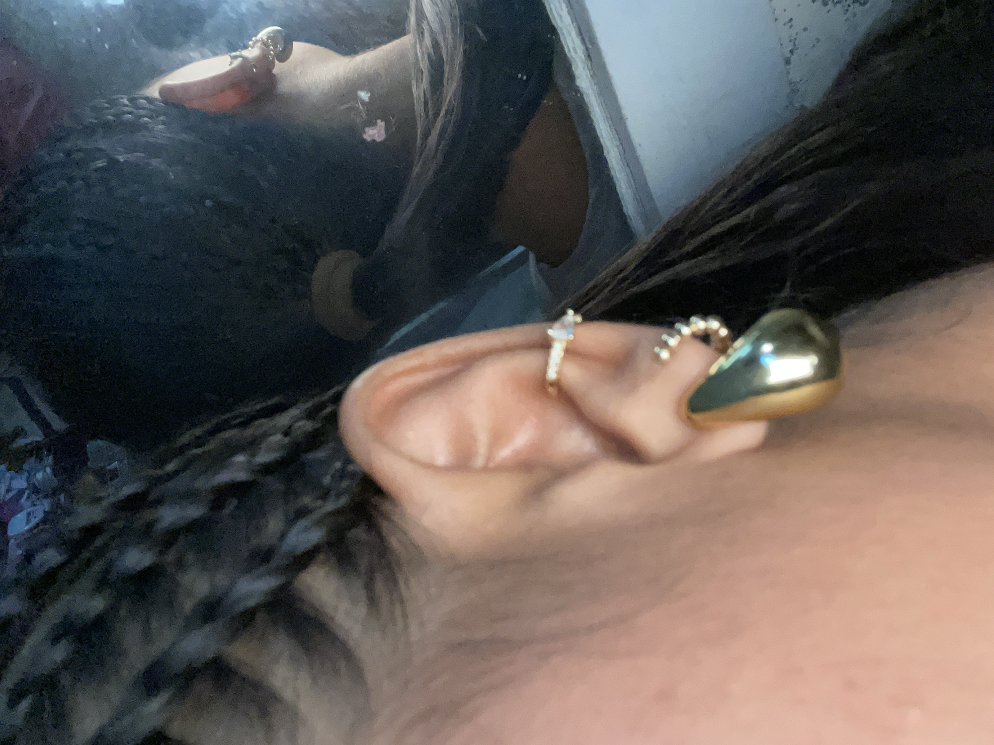 A close-up shot features a person's ear adorned with multiple earrings. The ear is pierced in three places, each holding a different earring: a simple ring, a gold beaded ring, and a larger gold dome-shaped earring. The skin appears smooth and the ear is well-defined. Dark braids are visible framing the ear and falling into the foreground. The background is blurred, suggesting a shallow depth of field. The location is Unknown, Unknown.