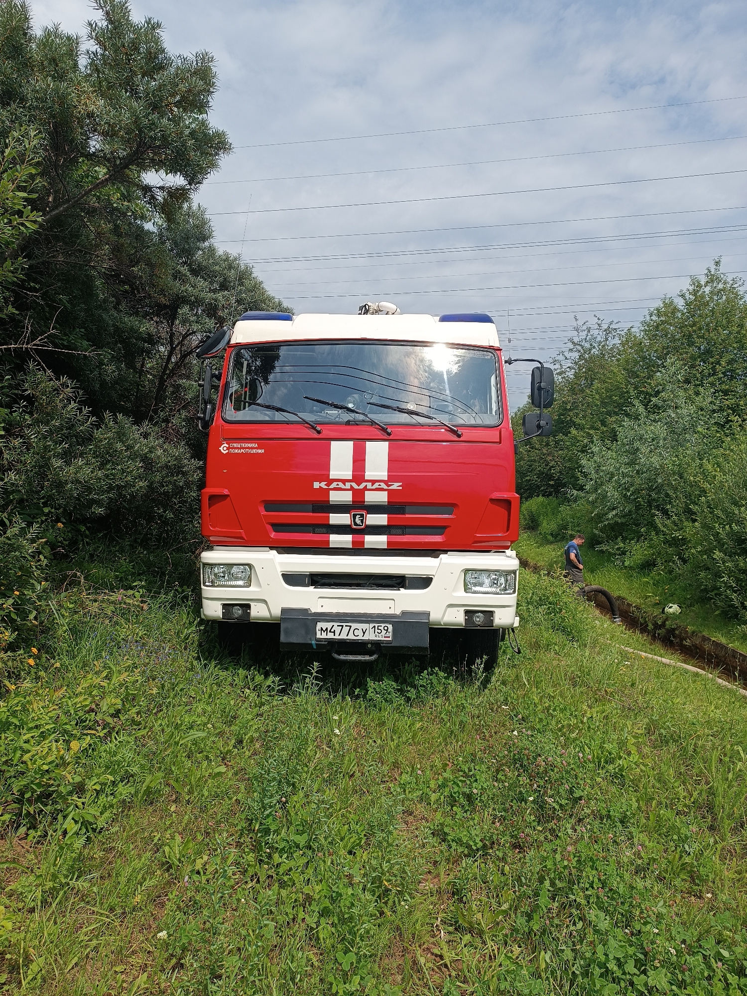 A red and white KAMAZ fire truck is parked on a grassy incline, partially obscured by trees. The truck has a blue and red light bar on its roof and "СПЕЦТЕХНИКА ПОЖАРОТУШЕНИЯ" (Special Equipment for Fire Fighting) written in Cyrillic on its side. A license plate with the number "M477CY 159" is visible. In the background, a person in a blue shirt is standing in a trench with a hose. The environment is overgrown with green grass and trees, under a cloudy sky with power lines running overhead.