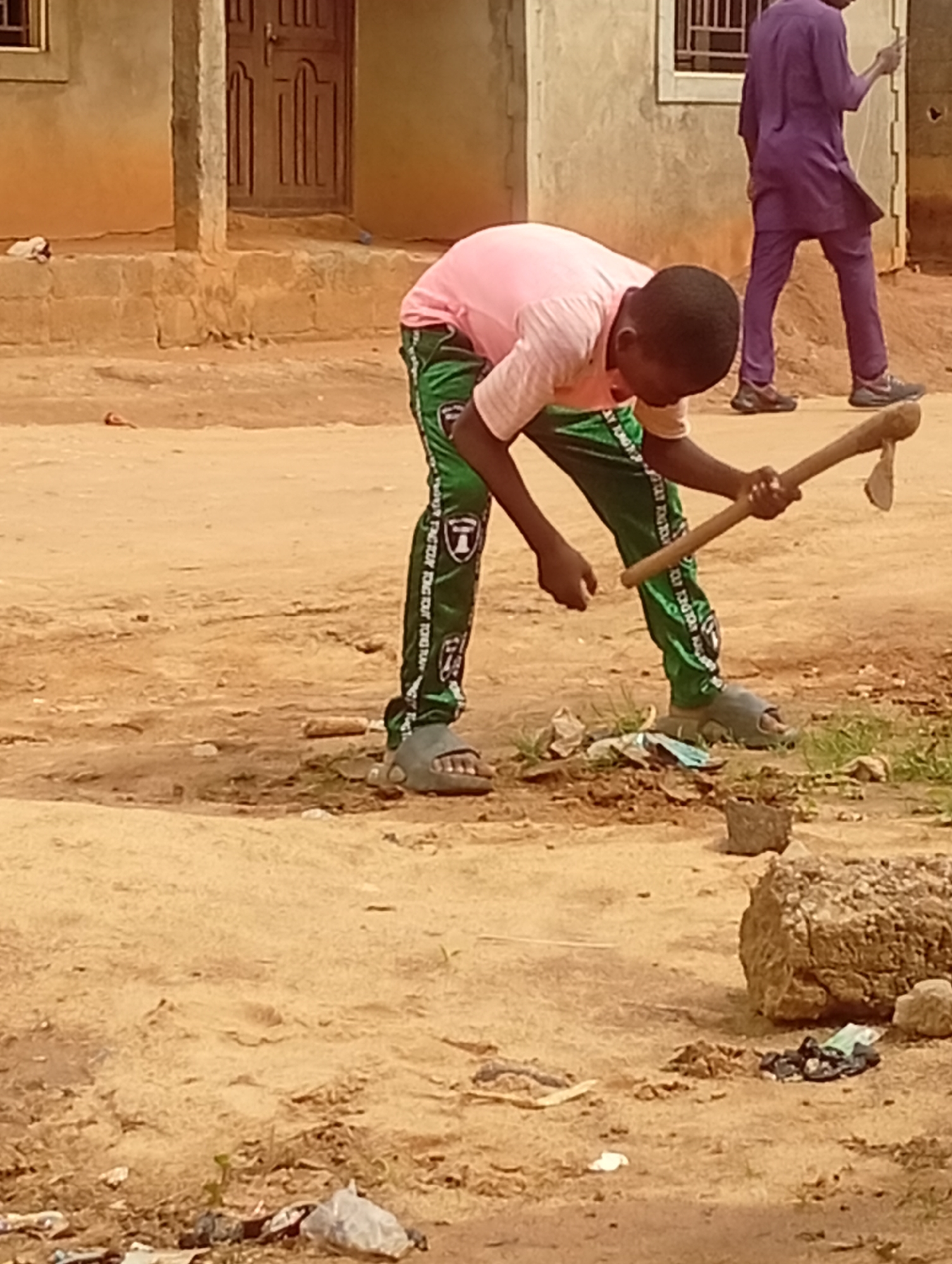 A young person wearing a pink shirt, green patterned pants, and grey sandals is standing on a dirt ground in Yelwa, Nigeria. They are holding an axe, apparently examining the blade's edge. The ground is littered with debris. In the background, there is a man in a purple outfit walking away. The buildings nearby are simple structures with brick walls.