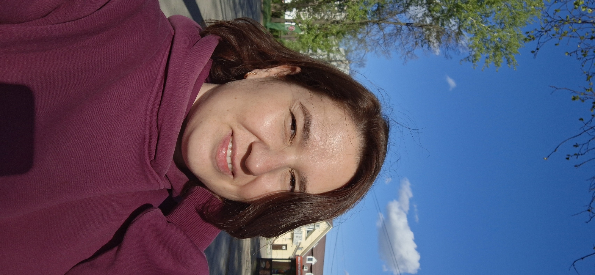 A woman is smiling, wearing a burgundy hoodie, with brown hair and fair skin. The picture is taken outdoors with a blue sky and some scattered clouds visible in the background. There is a building behind the woman and some trees, the location seems to be in the city of Chaikovsky, Russia.