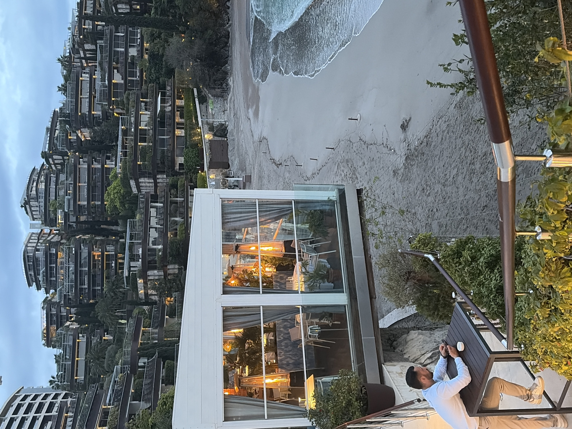 The image shows a view overlooking the beach in Budva, Montenegro, with the Dukley Hotel & Resort visible in the background. The beach is sandy, with the tide coming in, and has some wooden stakes in the sand. There is a modern, glass-enclosed structure, likely a restaurant or bar, with heating lamps inside. A man is leaning on a table on a balcony or patio, looking at a mobile phone. He is wearing a light-colored long-sleeved shirt and light-colored pants. A coffee cup is on the table. The architecture in the background is comprised of multi-level residences with terraces and vegetation. It is evening, with a slight glow visible on the beach.