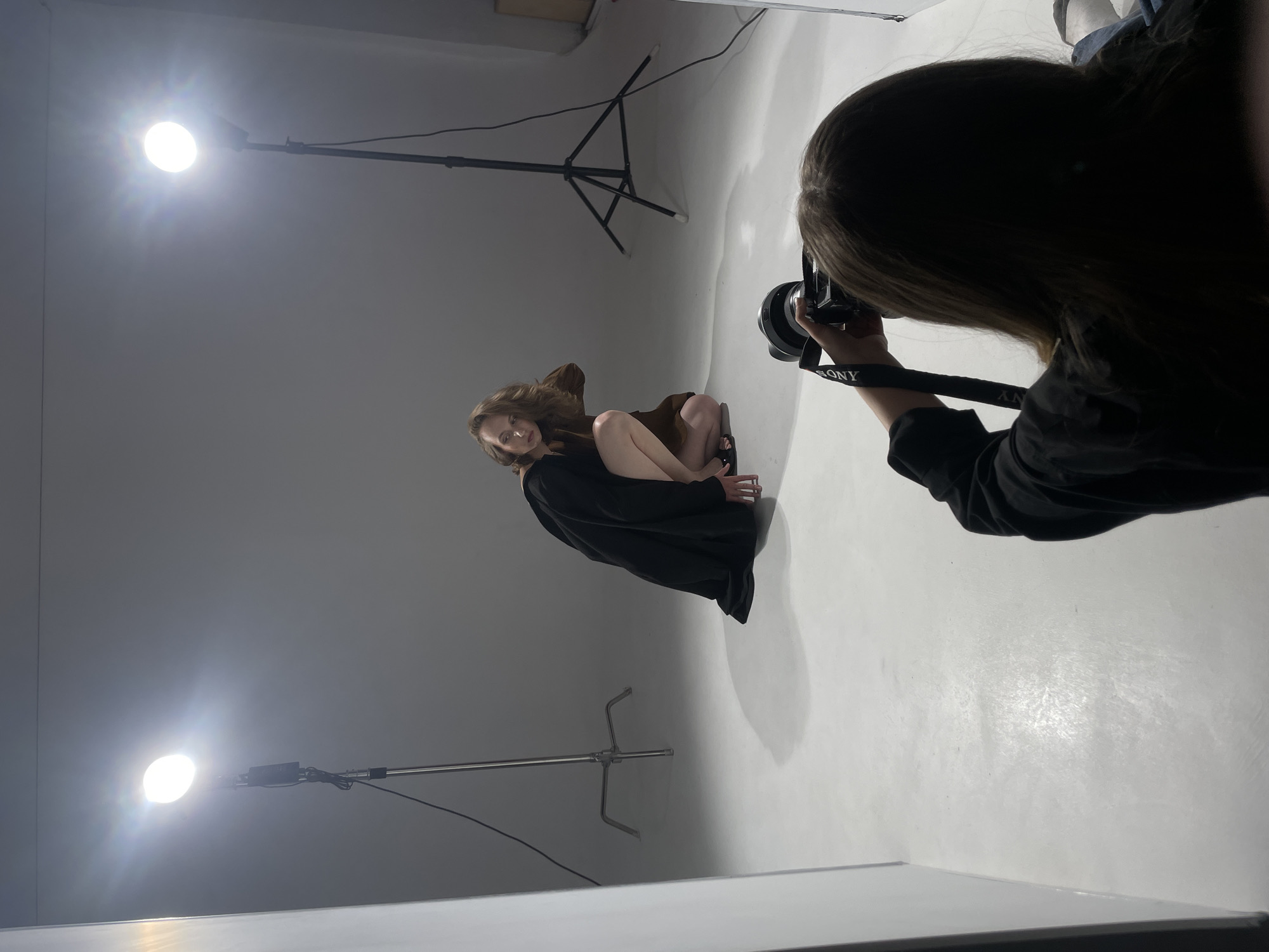 The image shows a photo shoot in a studio, likely indoors, not pinpointable to a specific location in Chaikovsky, Russia, without further evidence. A woman is posing, wearing a brown garment and a black robe or blanket draped around her. A photographer is positioned to the right, taking a picture with a Sony camera. The studio has two light stands with bright lights. The environment includes a white background and floor. The photographer is holding the camera and appears to be focused on capturing the image of the model.