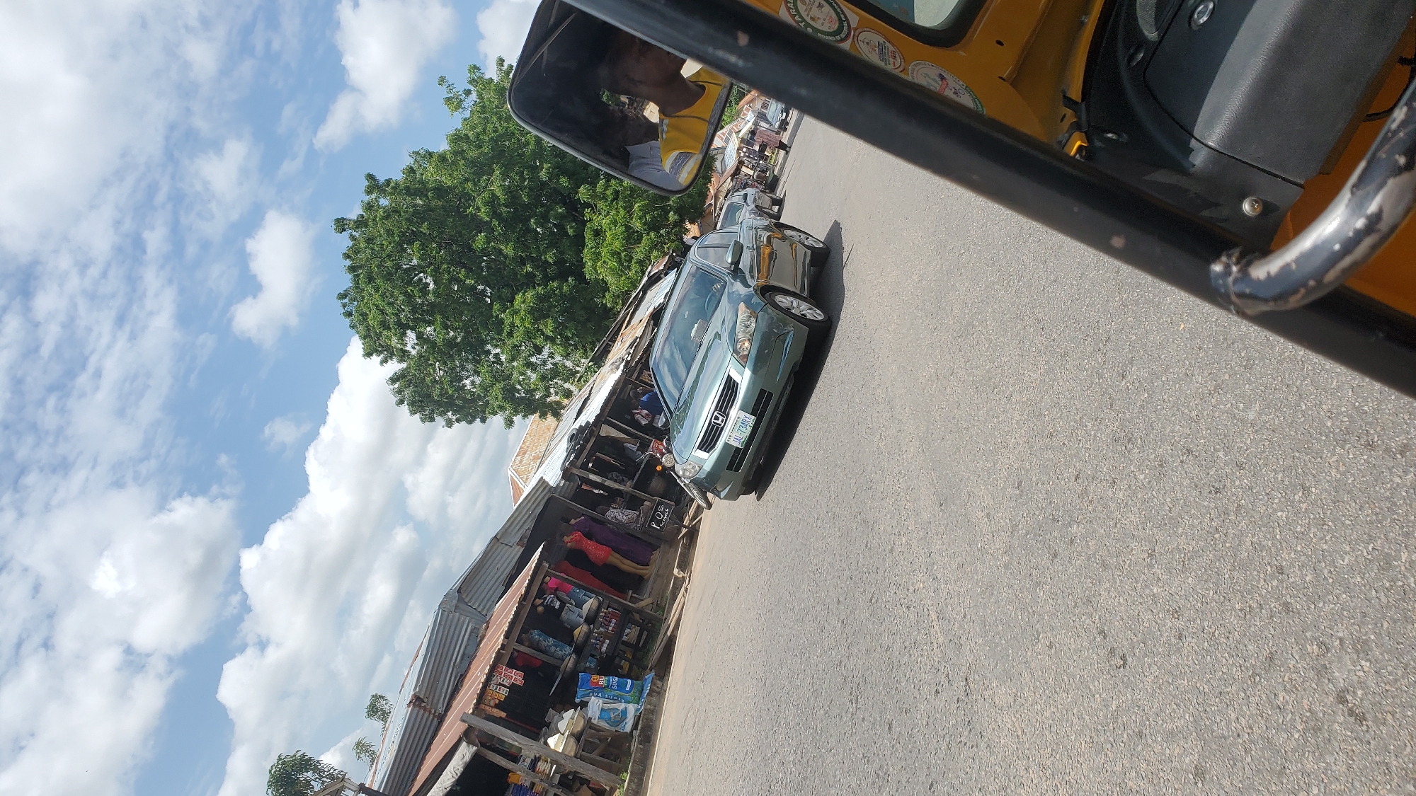 The image depicts a street scene in Jalingo, Nigeria. In the foreground, the yellow frame of a motorized tricycle is visible, with a portion of its dashboard and a side mirror. The side mirror reflects a person wearing a yellow shirt, presumably the driver or passenger of the tricycle.
On the road, a green sedan is driving away from the camera. The car has a license plate that reads "JAL-733ARX". To the left of the road, a row of open-air market stalls with corrugated metal roofs are set up. Various goods are displayed on tables and shelves, and several people are seen browsing or working at the stalls. One stall displays clothing.
Further down the road, more market stalls and buildings are visible. A large green tree stands behind the stalls, with a bright blue sky dotted with white clouds above. The overall impression is that of a bustling market area on a sunny day.