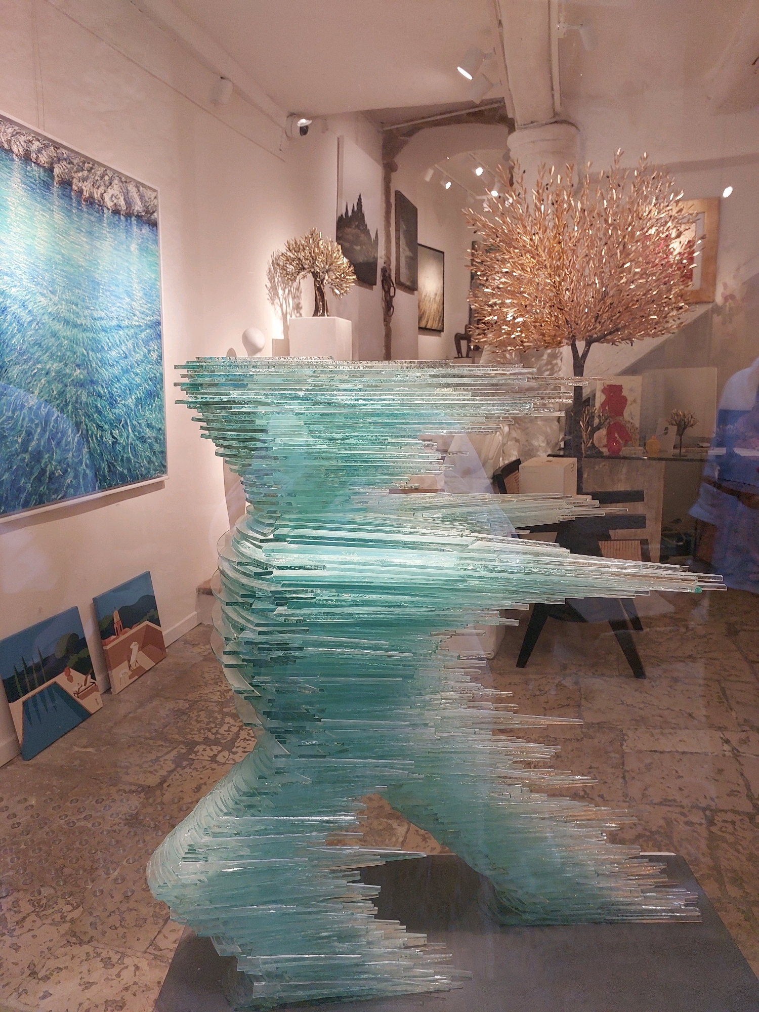 The image depicts an art gallery in Saint-Paul-de-Vence, France. A large, translucent sculpture made of layered glass, appearing as a stylized human form in motion, dominates the foreground. Behind the sculpture, various artworks are displayed on the white walls. To the left, a large abstract painting with vibrant blue and white hues hangs. Below it, two smaller rectangular paintings feature landscapes with buildings and figures. Further in the background, a metallic tree sculpture with golden leaves stands on a pedestal, illuminated by spotlights. Other paintings and sculptures are visible in the mid-ground and background, some reflecting in the glass sculpture. The floor is a patterned tile. The lighting suggests an indoor environment with artificial illumination, characteristic of an art exhibition space. There are no people clearly visible in the scene.