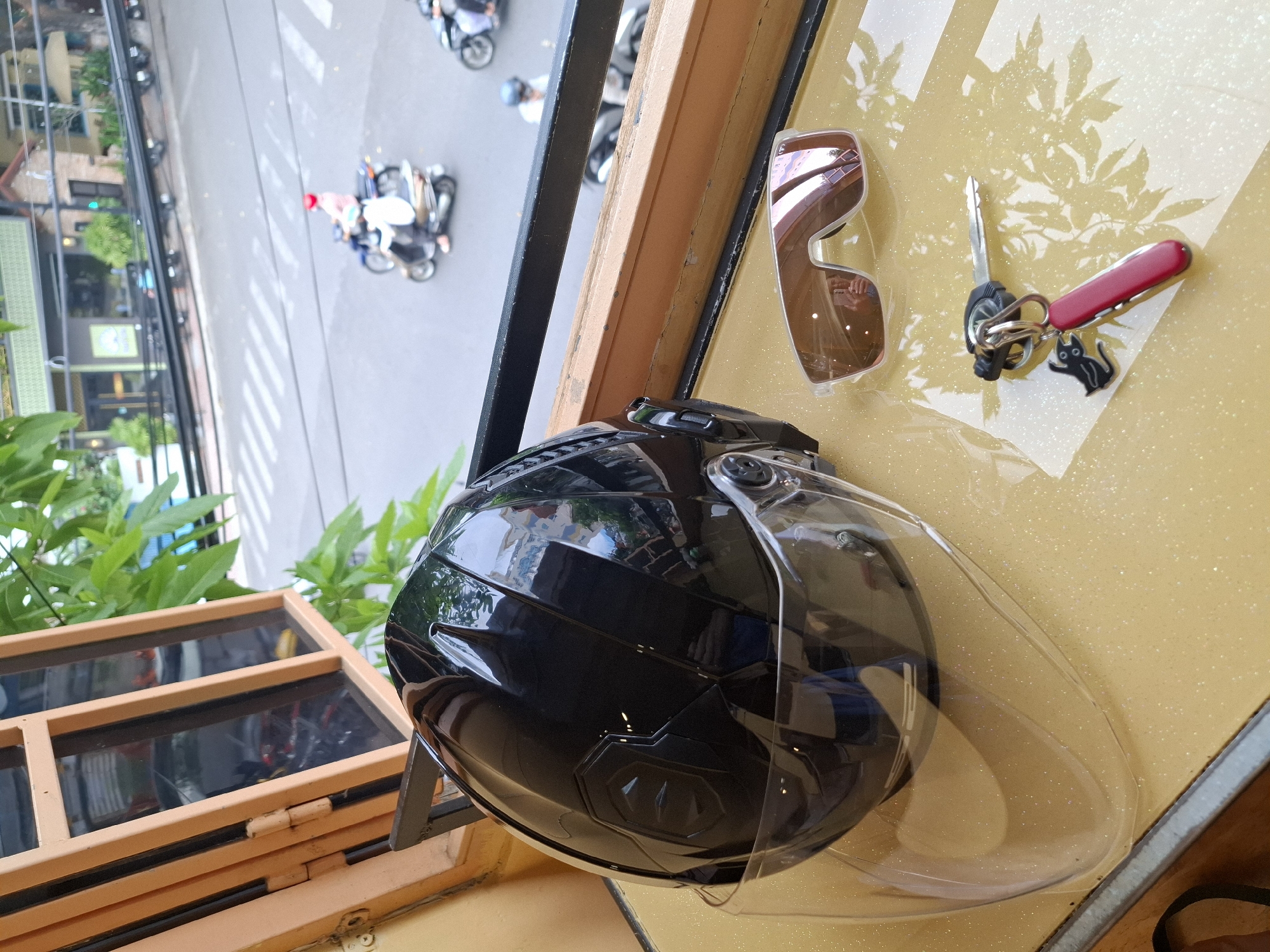 The image shows a black motorcycle helmet with a clear visor, a pair of sunglasses, and a keychain with a Swiss Army knife and a small black cat charm, resting on a yellowish surface. In the background, a street scene in Nha Trang, Vietnam is visible, with motorbikes and pedestrians. The scene appears to be captured from a slightly elevated perspective, possibly from a window or balcony, looking out onto a busy street. The overall impression is one of everyday life in a Vietnamese city, with a focus on personal items associated with motorbike travel.