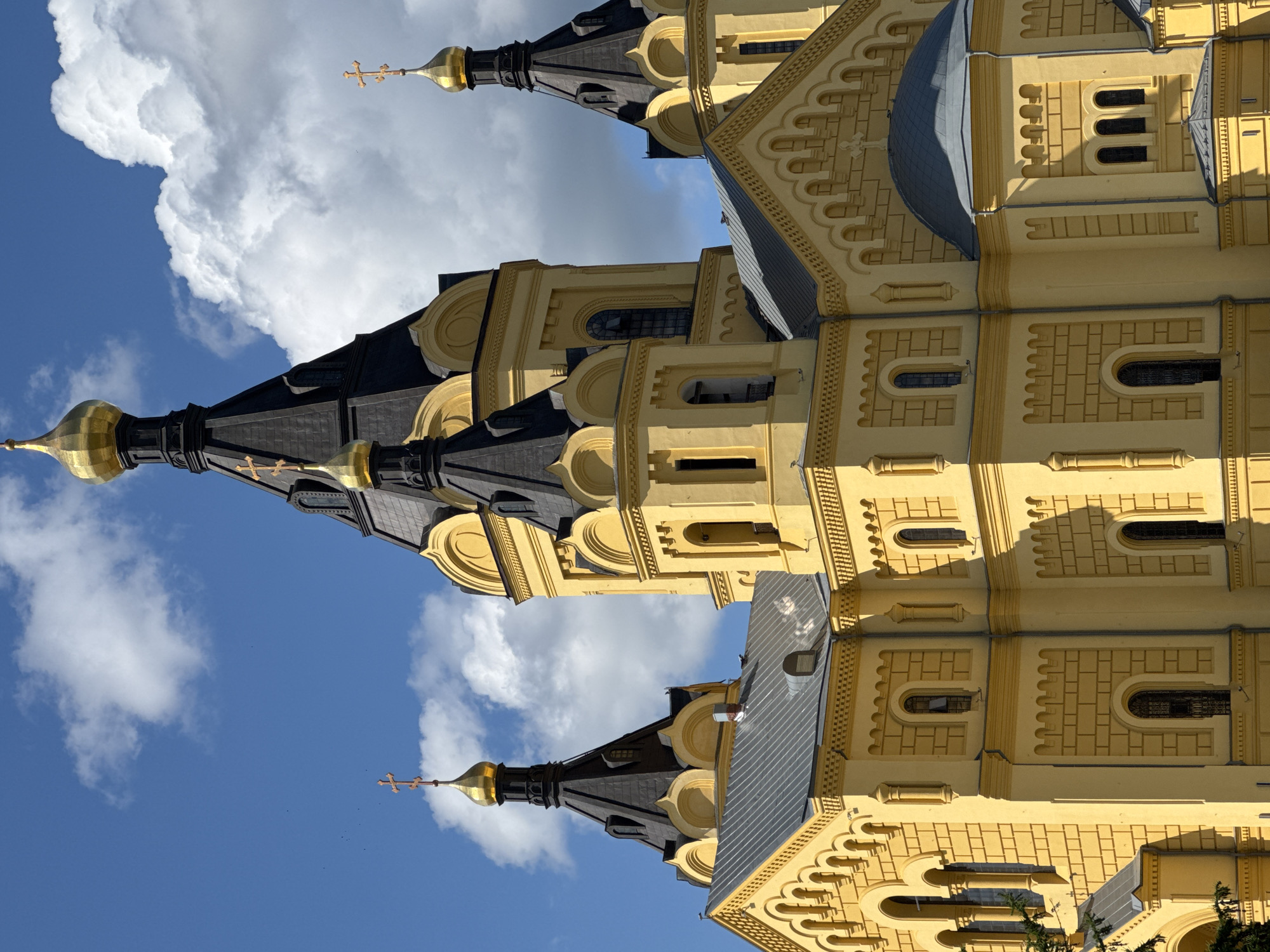 This is a close-up, eye-level, outdoor, medium shot of a large, yellow Orthodox church with multiple black and gold domes, set against a bright blue sky with white, puffy clouds. The architecture is ornate, featuring arched windows, decorative brickwork patterns, and numerous golden crosses atop the domes and spires. The sunlight casts strong shadows on the building, highlighting its textured facade. The image appears to be taken on a clear, sunny day. This is the Alexander Nevsky Cathedral in Nizhny Novgorod, Russia.