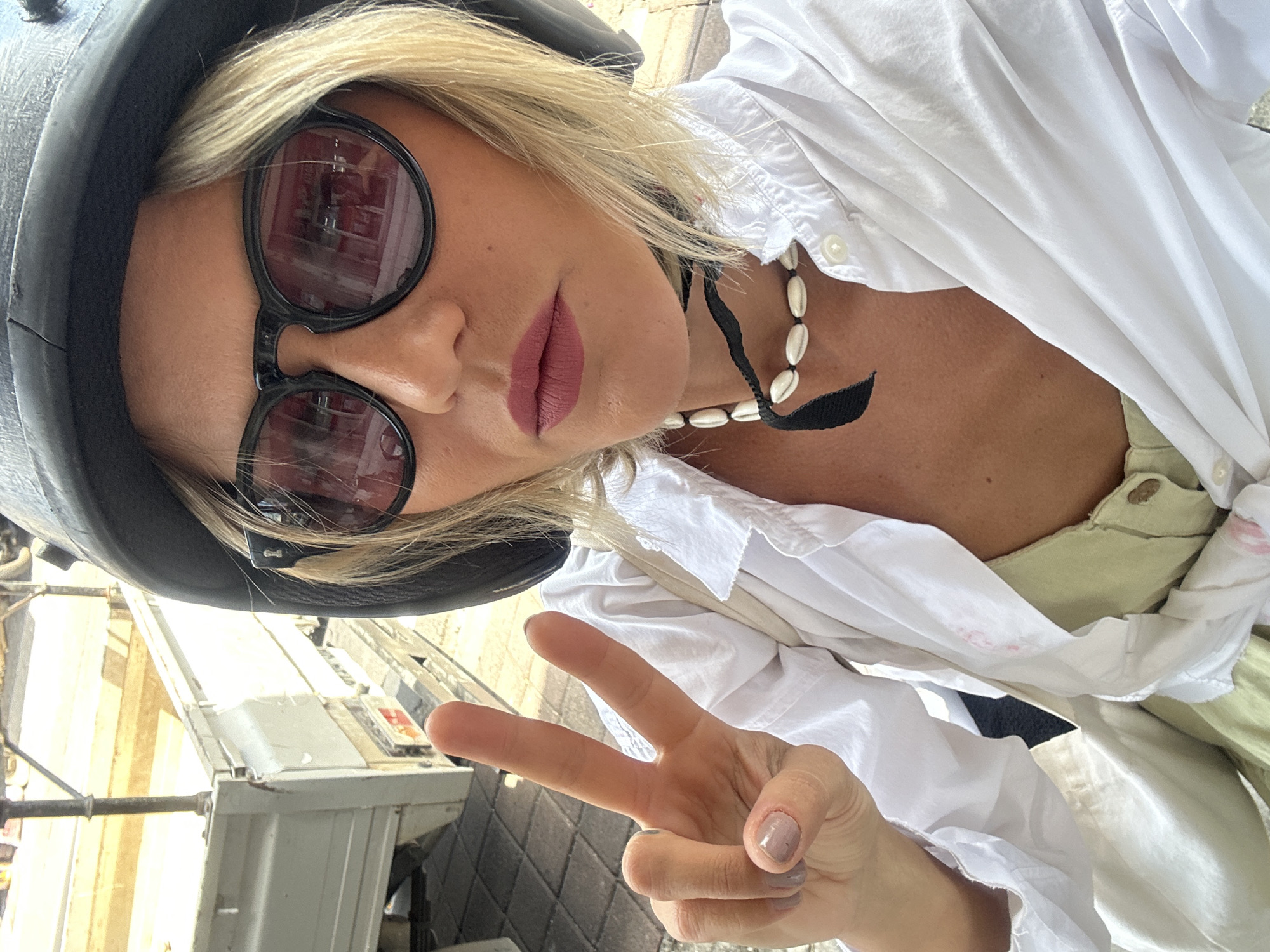 A woman with blonde hair and sunglasses wearing a black helmet, a white shirt, and a shell necklace poses for a selfie. She is holding up a peace sign with her right hand. The background is blurry and shows what appears to be a city scene, possibly North Kuta, Indonesia. A metallic structure is partially visible in the lower left. The photograph is rotated slightly counter-clockwise.