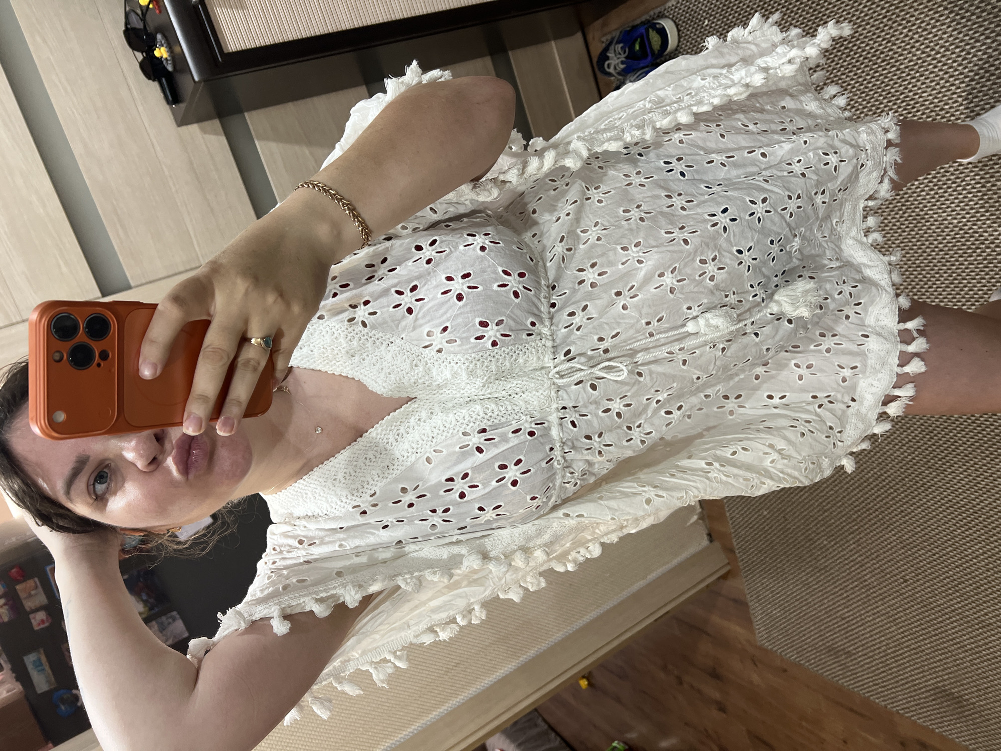 A woman is taking a mirror selfie in what appears to be a room. She is wearing a white, short, loose-fitting dress with intricate cut-out patterns and fringed trim. The dress has a V-neck and a drawstring waist. Underneath the white dress, a darker garment is visible. She is holding an orange smartphone in her right hand, capturing the reflection. Her left arm is raised, with her hand resting on her head. She is wearing a gold bracelet on her right wrist and a ring on her left hand. The background includes a wooden wardrobe with doors, a carpeted floor, and a partially visible doorframe. The scene suggests an indoor domestic setting, possibly a bedroom or dressing area. The location is identified as Noviy, Russia.