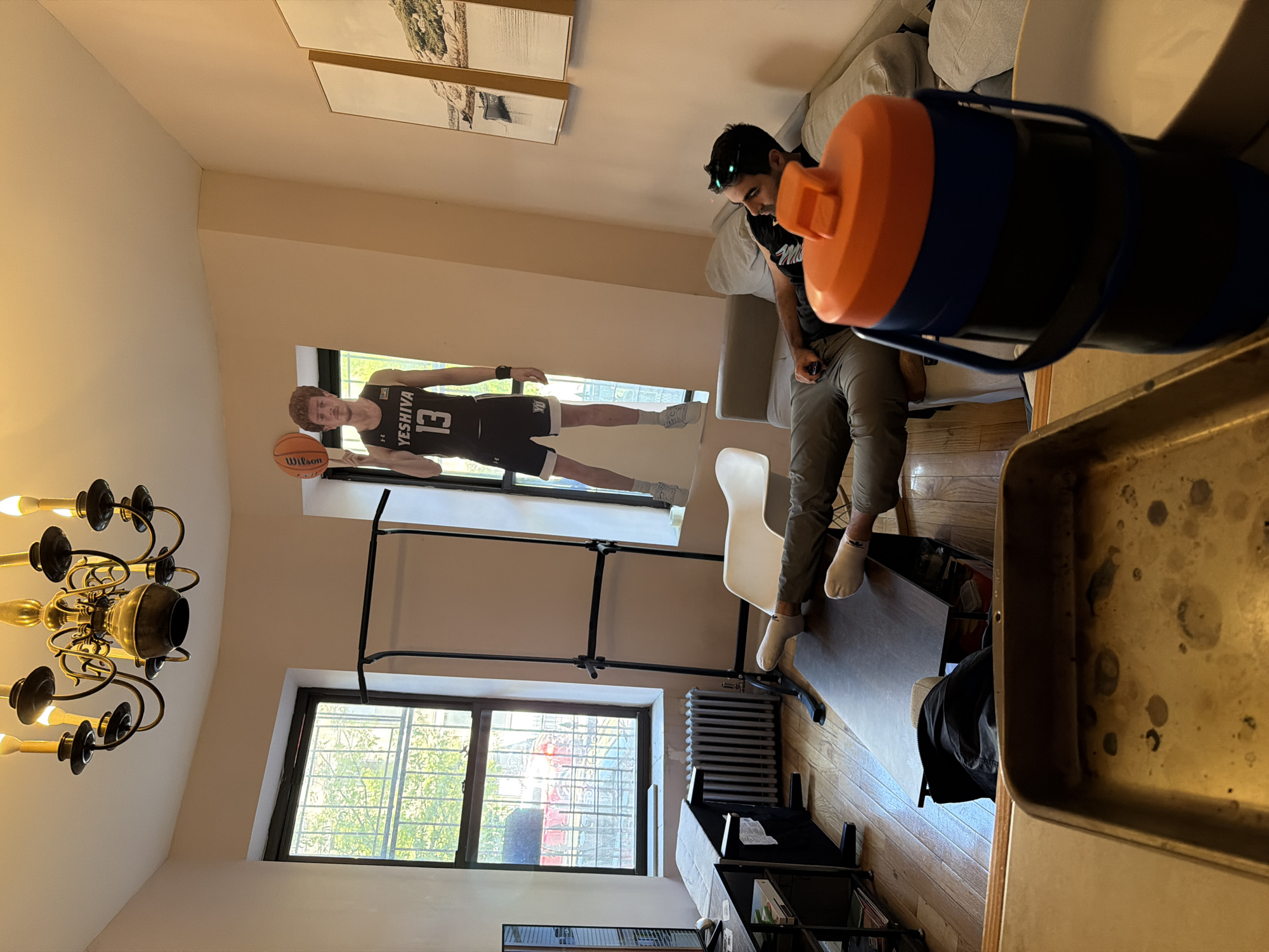 Indoor scene in a New York City apartment. A man sits on a chair, facing a pull-up bar mounted in a window frame, on which a life-size poster of a basketball player in a Yeshiva jersey is affixed. Outside the window, the urban landscape is visible through a barred window. The room includes a chandelier, a radiator, a sofa, and shelves. A blue and orange water bottle is in the foreground. A baking tray is in the lower right corner.