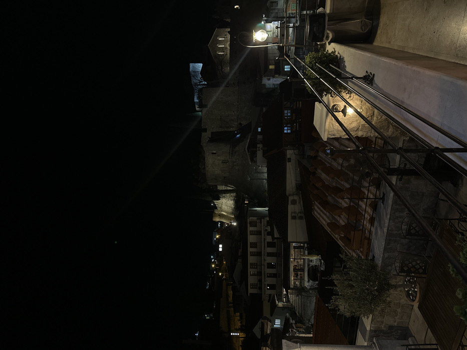 The image displays an elevated nocturnal view of Bartula, Montenegro. In the foreground, a terrace or balcony is defined by a dark metal railing with spherical finials and stone pillars. An illuminated wall sconce is affixed to a central stone pillar, casting light on adjacent green foliage. To the left, a potted olive tree stands near a partially visible wooden table and dark metal chairs. A tall, decorative lamp post with a spherical globe is positioned on the far right of the terrace. Below, numerous multi-story buildings with red-tiled roofs descend into the town, some exhibiting illuminated windows. An outdoor dining area featuring tables, chairs, and a striped awning is visible on one of the mid-level structures. In the background, a large, fortified stone structure, consistent with a castle or fortress, dominates a prominent hillside, illuminated by external spotlights. A secondary stone building with arched openings is situated to the fortress's right, also illuminated. The sky is dark, indicating night, with scattered artificial lights visible across the distant landscape. No individuals are present in the scene, and no overt actions or interactions are depicted.