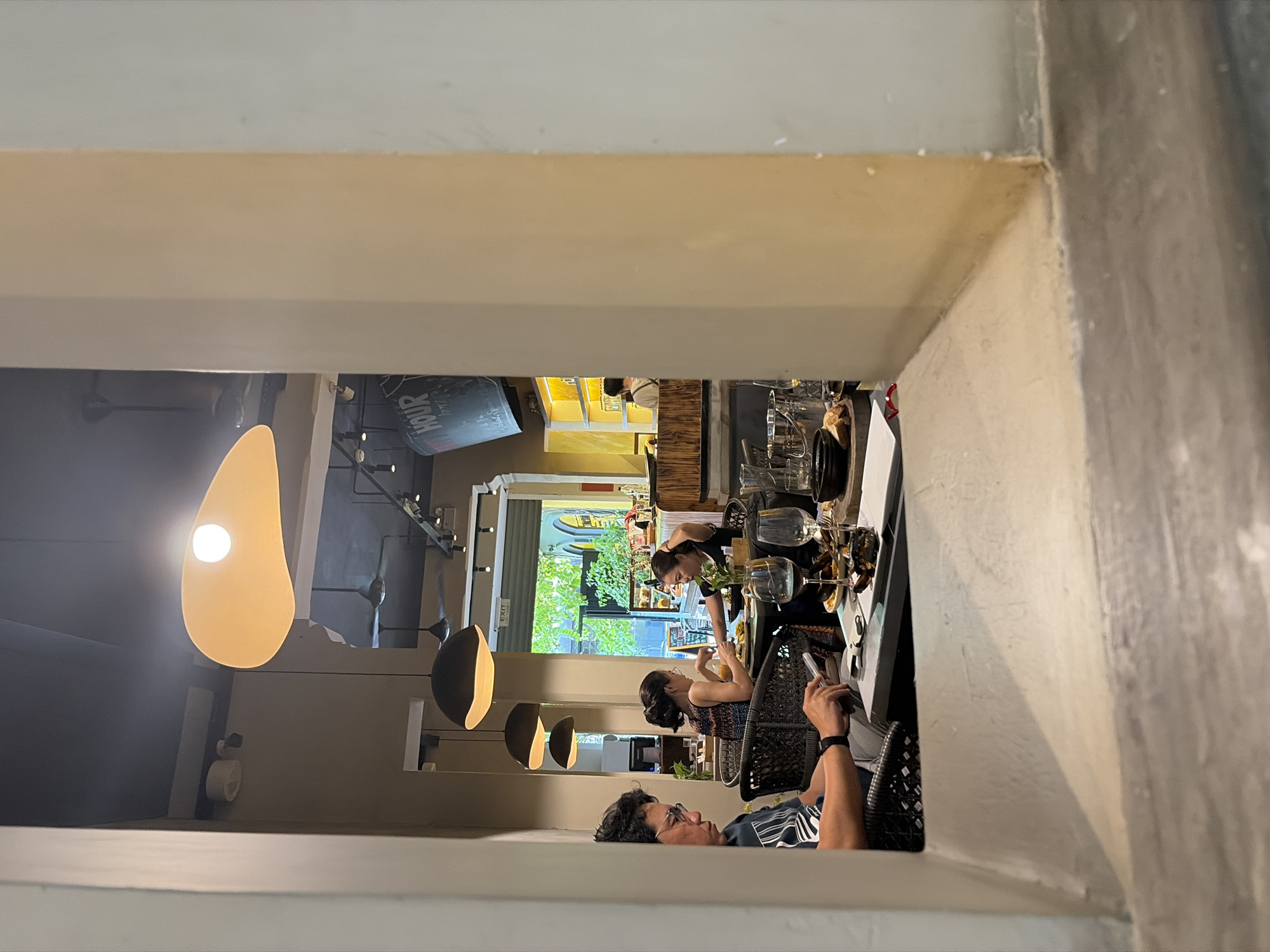 The image depicts an interior view of a cafe or restaurant, framed by a beige architectural opening, likely in Thủ Đức, Vietnam.
In the foreground, a prominent beige-colored architectural element, possibly a pillar or wall segment, occupies the left and right sides of the frame, with a rough-textured ledge forming the bottom.
Beyond the frame, the cafe interior is visible. On the left, a man with dark, curly hair and glasses, wearing a dark striped t-shirt and a smartwatch, is seated at a table, focused on a silver smartphone held in both hands. His chair is dark and woven. Behind him and slightly to the right, two women are seated at another dark table. One woman, with dark hair tied back and wearing a patterned top, holds a glass of orange liquid. The other woman, partially obscured, has dark hair and a dark top, appearing to be dining.
The table in the mid-right features two empty wine glasses (one with red residue), a plate holding dark, possibly grilled, food items, a small wooden board, and a silver bucket with other drinkware. The ceiling is dark grey, from which three dark, conical pendant lights hang, along with one large, irregular yellow-orange pendant light providing central illumination. Two dark ceiling fans are also visible.
In the background, an open arched doorway reveals an exterior scene with vibrant green foliage, indicative of an urban street. An "EXIT" sign is mounted above this doorway. To the right of the exit, a bar area is visible with a staff member, seen from the back, attending to it. Behind the bar are brightly illuminated yellow shelves displaying bottles and glassware. A large, dark sign with the word "HOUR" in white lettering is partially visible in the upper right background. The overall lighting is warm, creating an inviting ambiance.
