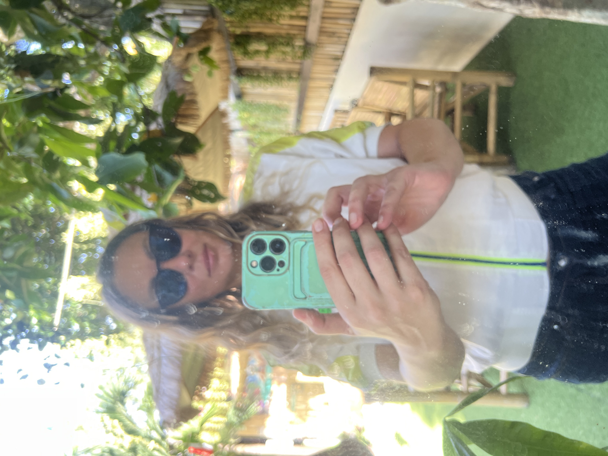 A person is taking a selfie in a mirror, holding a green smartphone. The person is wearing sunglasses, a white jacket with neon green accents, and dark pants. The reflection shows a tropical outdoor setting with lush green foliage, thatched-roof umbrellas, and bamboo fencing. In the background, there are bamboo chairs and a grass-like floor. The lighting suggests it is daytime, with sunlight filtering through the leaves. The specific location in ตำบลเกาะพะงัน, Thailand, appears to be an outdoor seating area, possibly at a resort or cafe, characterized by its tropical ambiance.