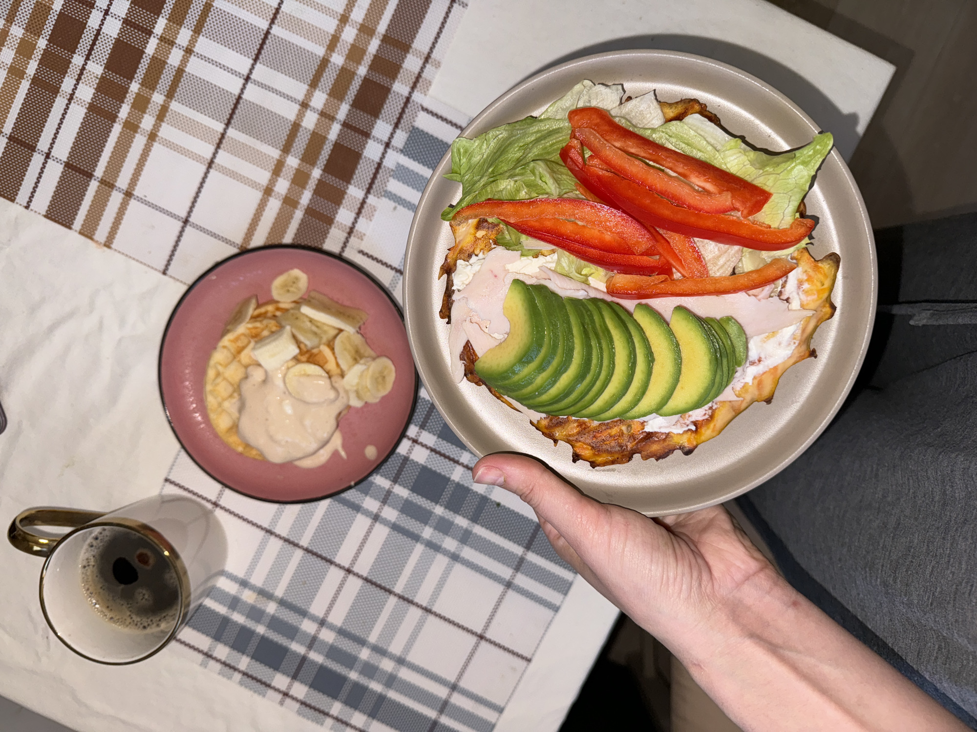 The image shows a person holding a plate of food, a pink plate with what appears to be a pancake and bananas, and a mug of coffee on a checkered tablecloth. The plate held by the person contains a circular, savory base topped with lettuce, sliced red bell peppers, sliced avocado, and some white filling. The person's hand and part of their arm are visible, wearing a grey long-sleeved shirt. The background shows a white surface and a checkered tablecloth with brown, tan, and white squares. The pink plate has a pancake or waffle topped with sliced bananas and a light-colored sauce. The coffee mug has a gold-colored handle and contains black coffee. The location in Krasnoyarsk, Russia is not determinable from the visual information provided.