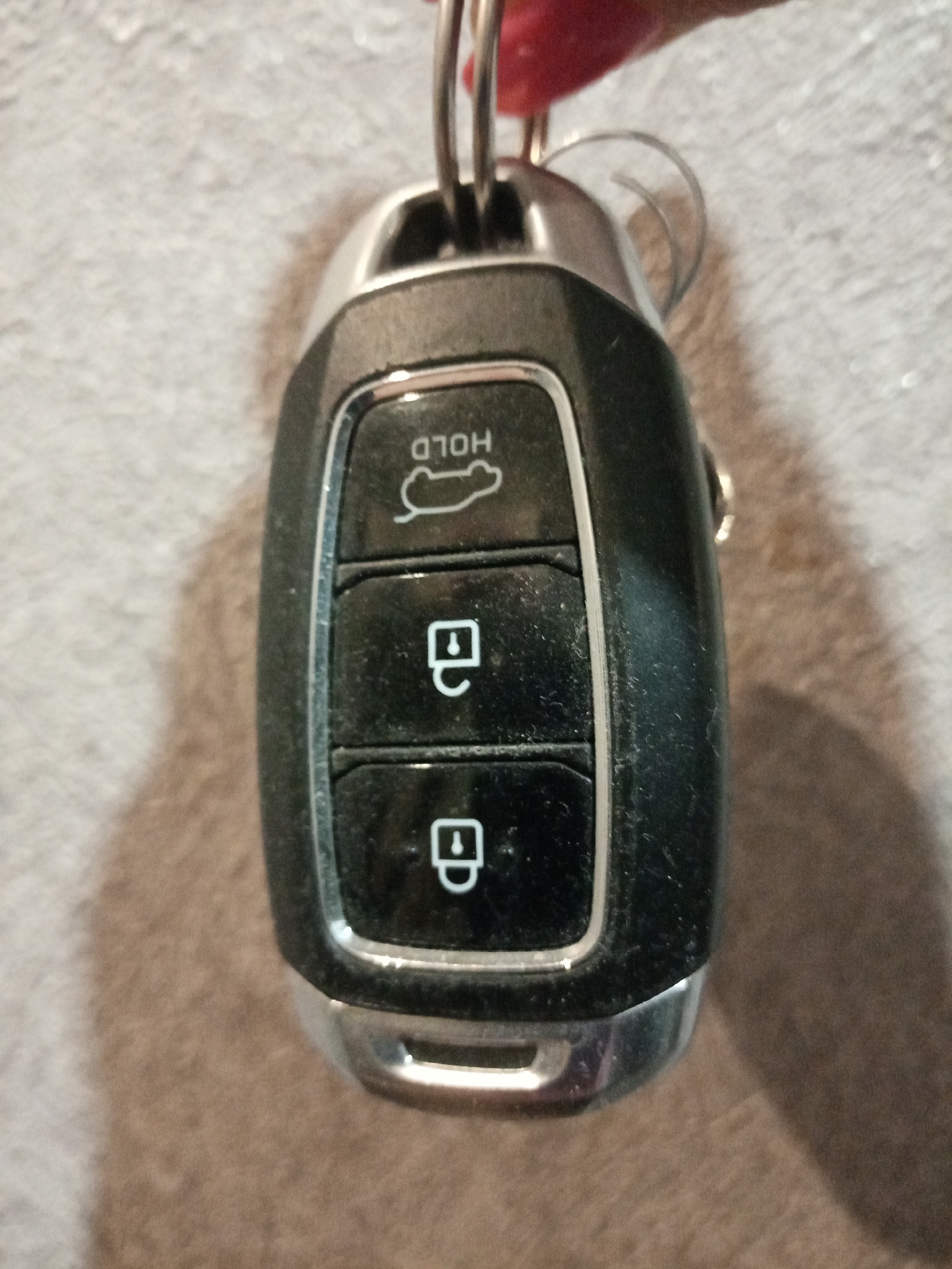 The image shows a car key held in someone's hand against a blurred background of what appears to be a carpet or fabric surface. The key is a modern, electronic car key with buttons for trunk release, lock, and unlock. The top button is labeled "HOLD" with a symbol of an open trunk. The next two buttons display a lock symbol, one indicating lock and the other, unlock. The key has a black body with a silver trim at the top and bottom. The keyring is visible. There is no information in the image to indicate the location is in Elektrostal, Russia.