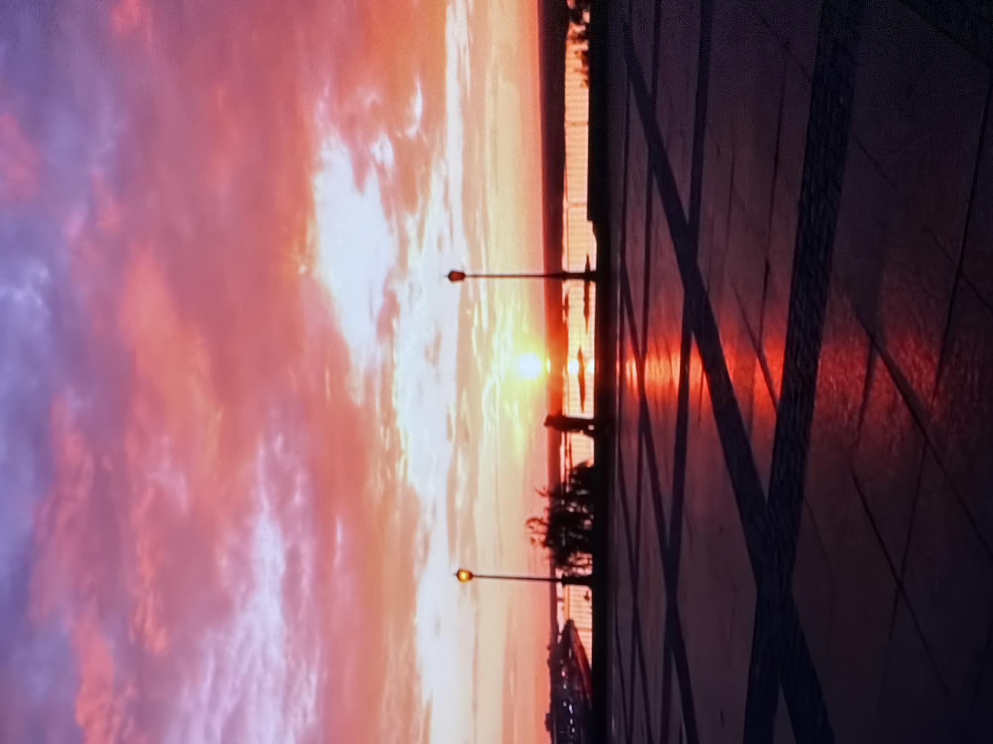 The image depicts a sunset over a body of water in an urban environment. The sky is filled with vibrant pink and orange clouds, illuminated by the setting sun. In the foreground, a paved plaza with a geometric pattern of dark and light tiles reflects the warm colors of the sunset. Two streetlights are visible, one with a lit lamp, positioned on the edge of the plaza overlooking the water. A silhouette of a person stands near the water's edge, and another person is partially visible in the distance. The water itself reflects the dramatic colors of the sky, creating a shimmering effect. The scene suggests a tranquil evening at a waterfront promenade.