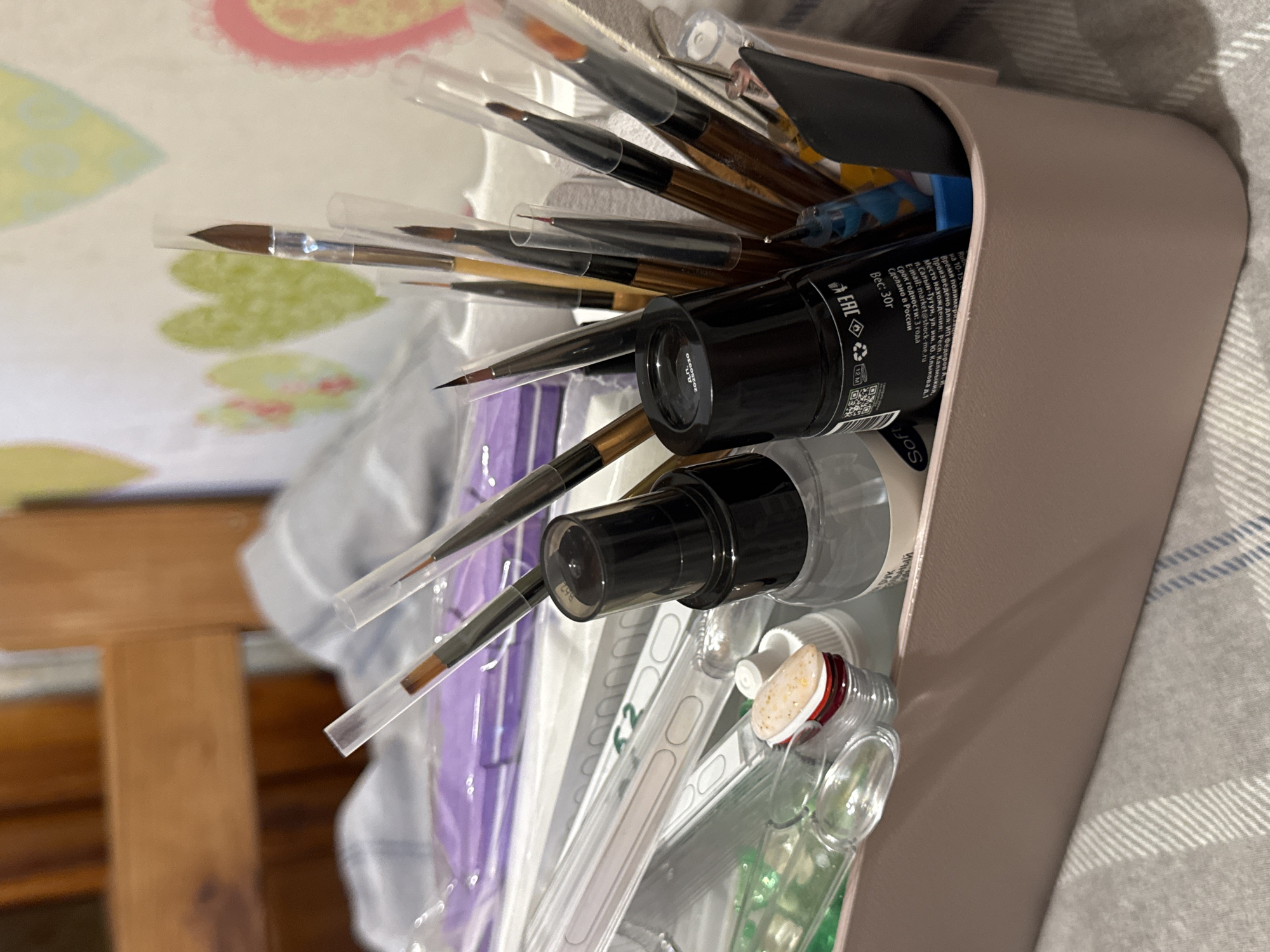 The image depicts a close-up view of a light pink organizer box filled with various nail art supplies. The organizer contains an assortment of brushes with clear protective sleeves, bottles of nail polish or gels, and other small tools. A black bottle with Russian text is prominently visible in the center. The background shows a wooden chair and a patterned cloth or curtain with heart designs. The overall impression is of an organized collection of items for nail art, likely in a personal space. The context of Яромінскі сельскі Савет, Belarus, is not visually discernible from the image itself.