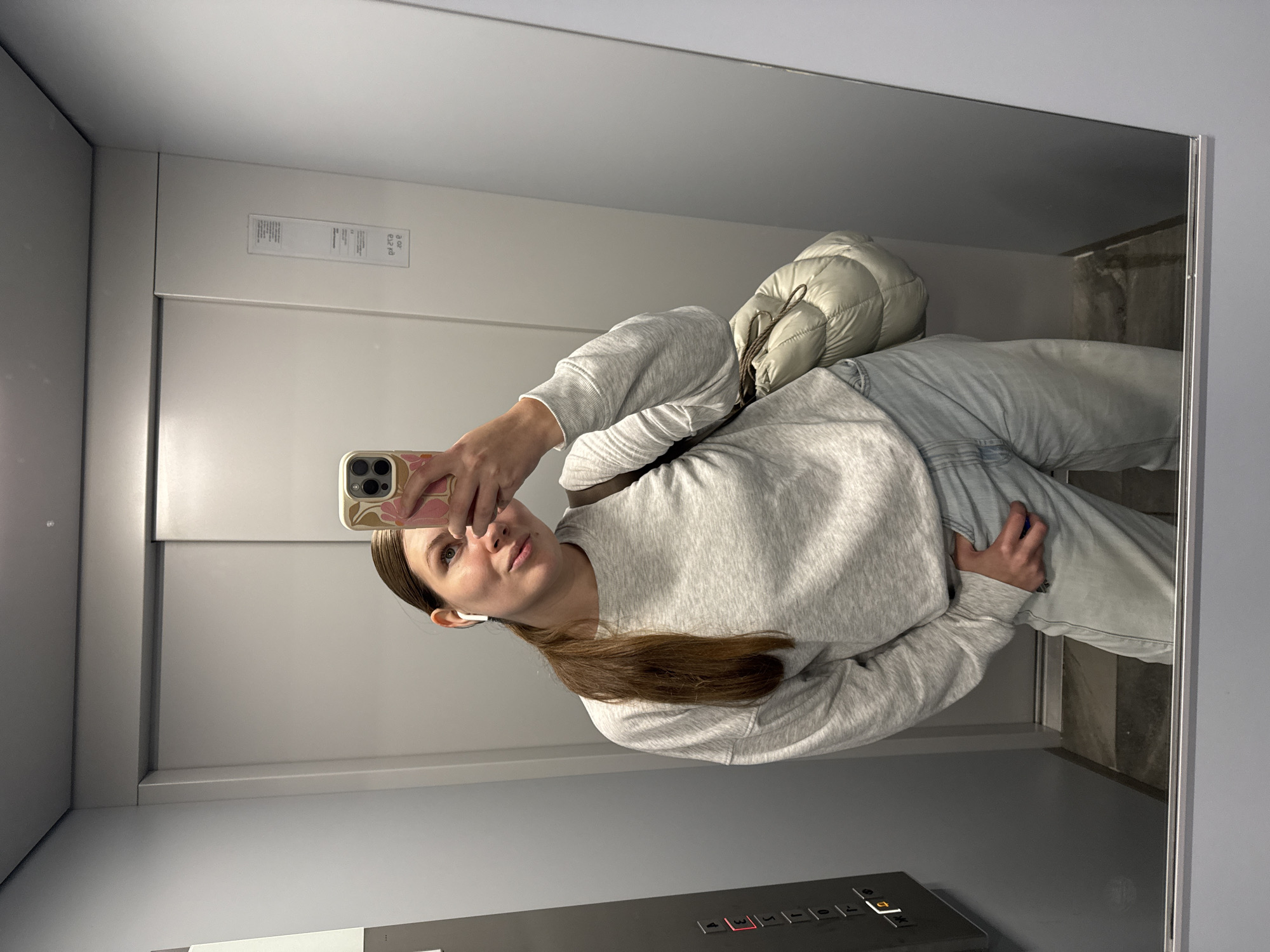 The image shows a woman taking a selfie in what appears to be an elevator. She is wearing a light gray sweater and jeans. A white, puffy bag is slung over her shoulder. The elevator has gray metal walls and a panel with buttons indicating floors, one of which is illuminated "3". The elevator appears to be in good condition and well-maintained. The image contains no visual references to determine that it was taken in Vozdovac Urban Municipality, Serbia.