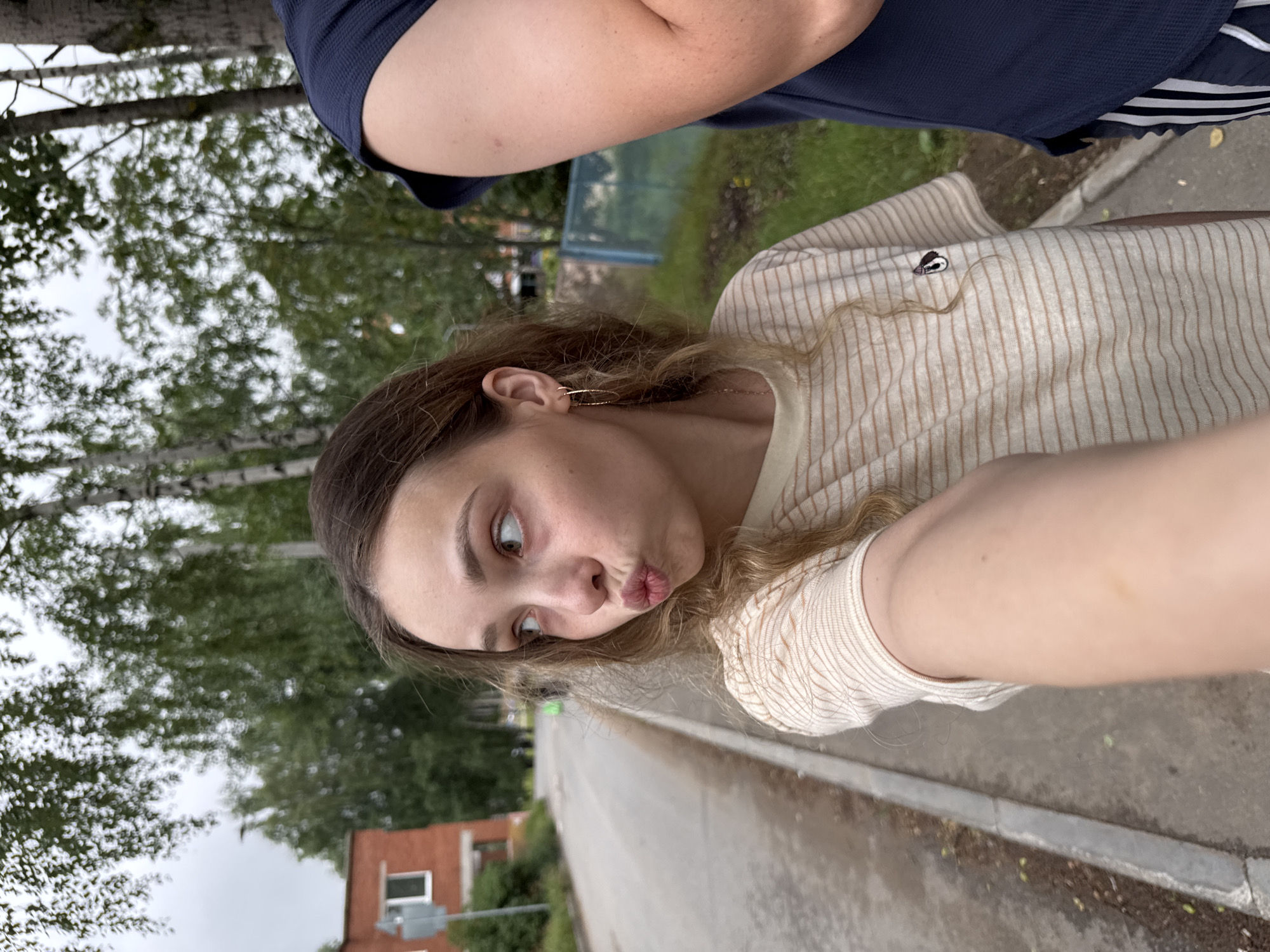 A young woman with long, wavy brown hair and light brown eyes takes a selfie outdoors. She is wearing a light-colored, striped shirt. Her face is turned towards the camera, and she is puckering her lips in a playful expression. Her right arm is visible, extended towards the camera. Behind her, a person wearing a dark blue shirt is partially visible. The background shows trees with green leaves, a paved walkway, and a brick building. The scene appears to be set in a suburban or residential area in Noviy, Russia. The lighting suggests it is daytime, possibly overcast due to the diffused light and muted colors.