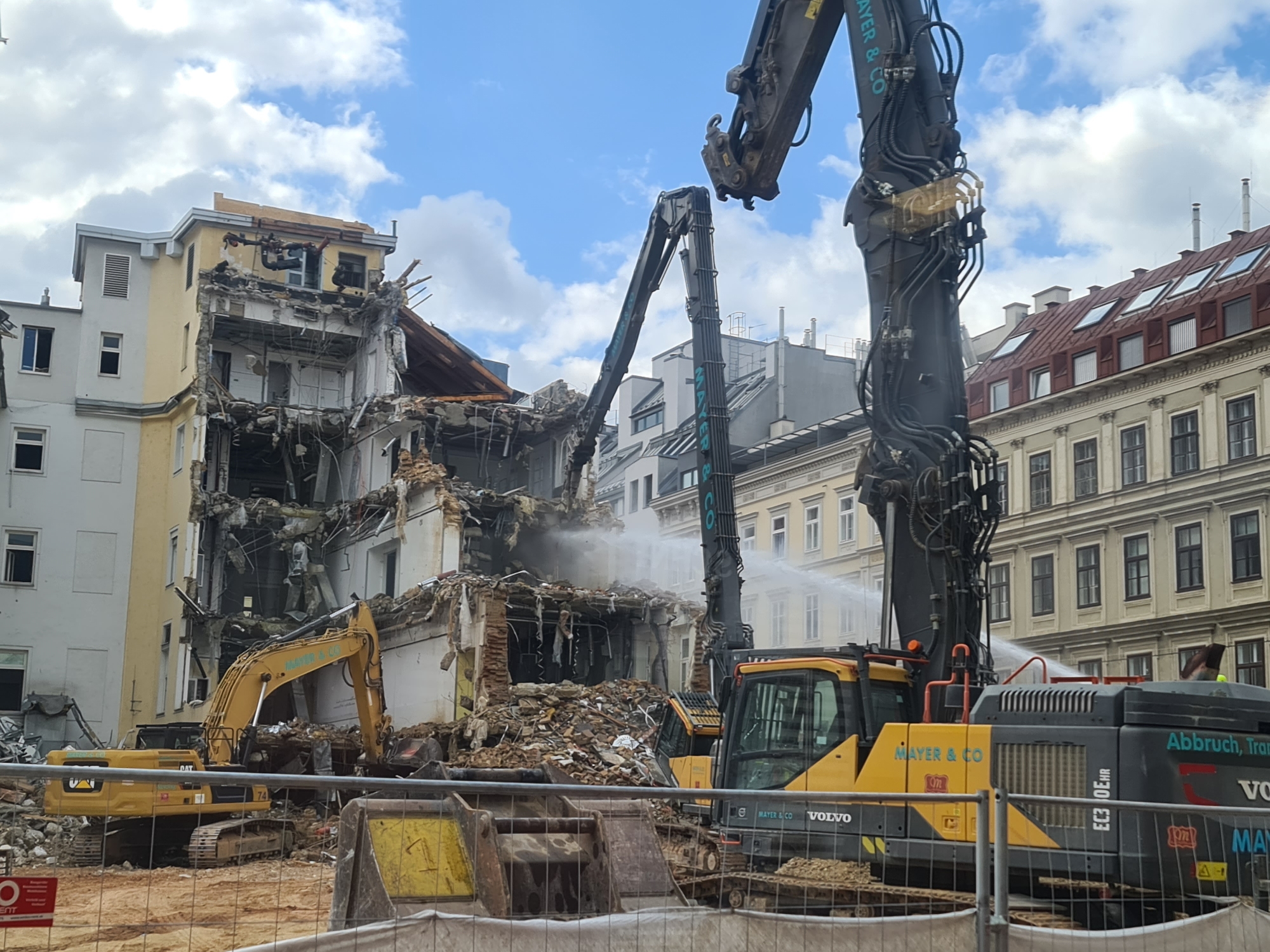 This image shows a demolition site in Vienna, Austria. In the foreground, two yellow excavators are visible. The excavator on the left has "CAT" and "74" printed on its side. The excavator on the right is a Volvo with "MAYER & CO" and "EC30EHR" printed on its body. A large, dark excavator arm with hydraulic hoses extends from the right side of the frame, pointing towards a partially demolished building. Water is being sprayed from above, likely to control dust.
The building in the center of the image is in a state of demolition, with debris scattered around its base. Sections of the building are missing, revealing its internal structure. The facade of the building is a mix of yellow and white. Behind the demolished building, other buildings with red roofs and many windows are visible, characteristic of Viennese architecture. The sky is blue with scattered white clouds. A safety fence is present in the foreground, separating the viewer from the demolition zone. A red and white sign with a symbol is attached to the fence on the left.