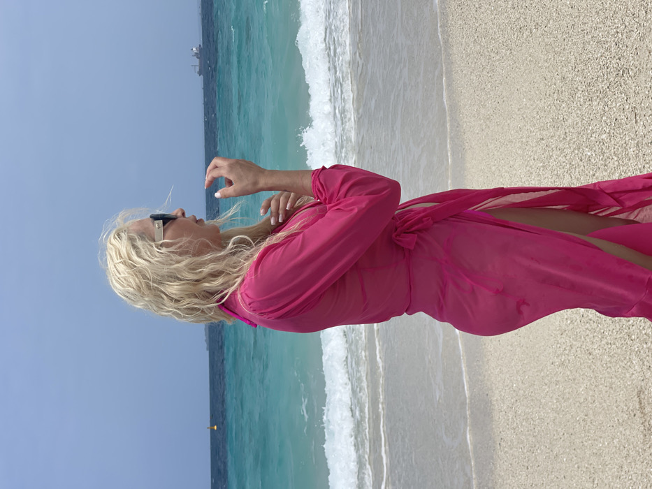 A blonde-haired female, wearing dark sunglasses and a sheer pink long-sleeved cover-up, stands in profile on a light-colored sandy beach in the city of Zanzibar, Tanzania. She faces right, looking towards the turquoise and dark blue ocean. Her left hand is raised near her chin, with fingers slightly curled. Gentle waves break on the shore directly behind her. In the middle distance to the left, a yellow buoy is visible in the water. On the far horizon to the right, two large cargo vessels are present under a light blue sky.
