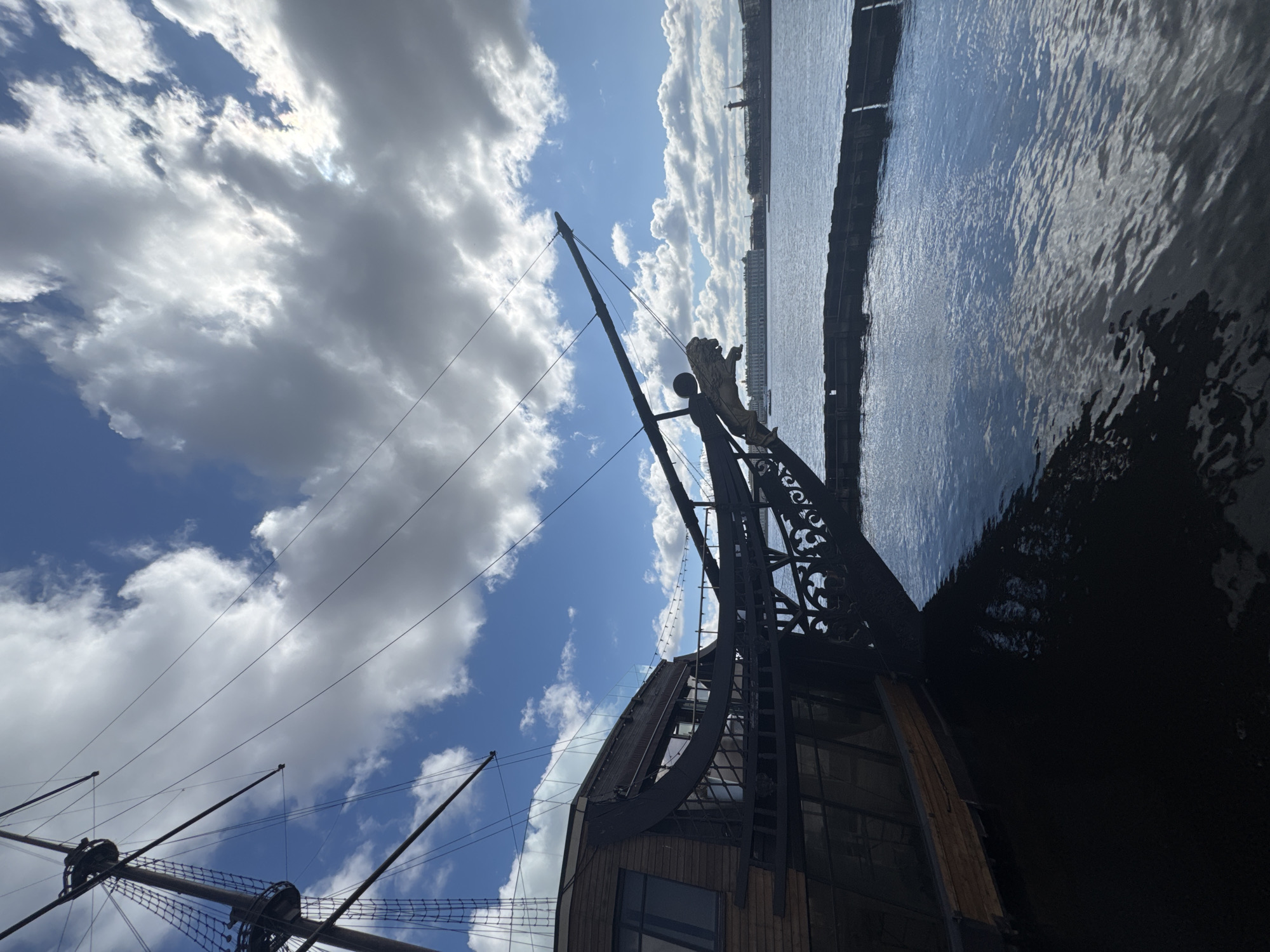 View of the frigate " Благодать " restaurant ship, in Saint Petersburg, Russia. The ship's decorative bow features a lion figure. The sky is mostly blue with white clouds. The Neva river is visible on the right.