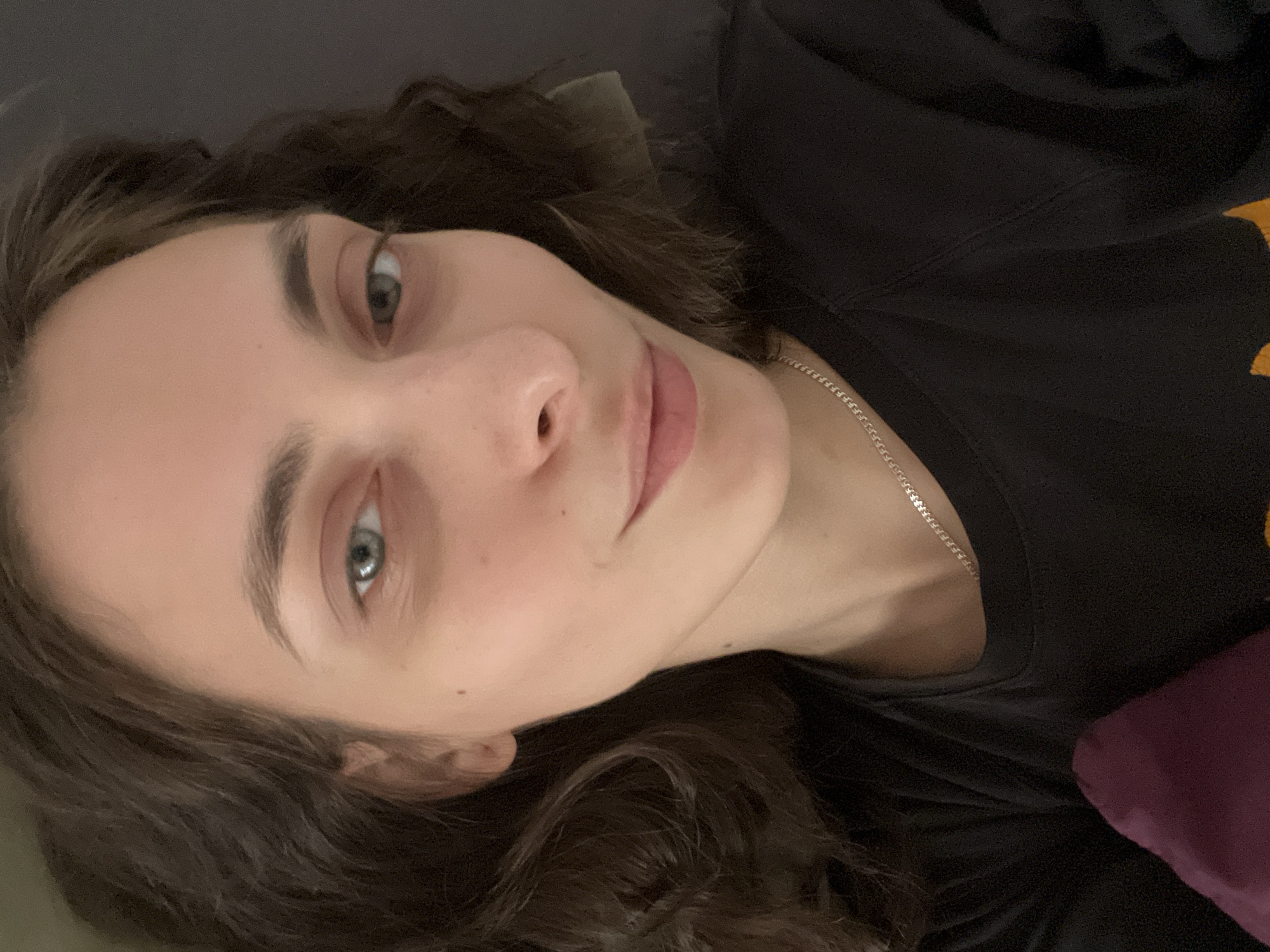 A young woman is lying down, facing the camera. She has dark hair and light eyes. She is wearing a black top and a silver necklace. The background is blurred and dark. There is no indication of this photo being taken in Krasnoyarsk, Russia.