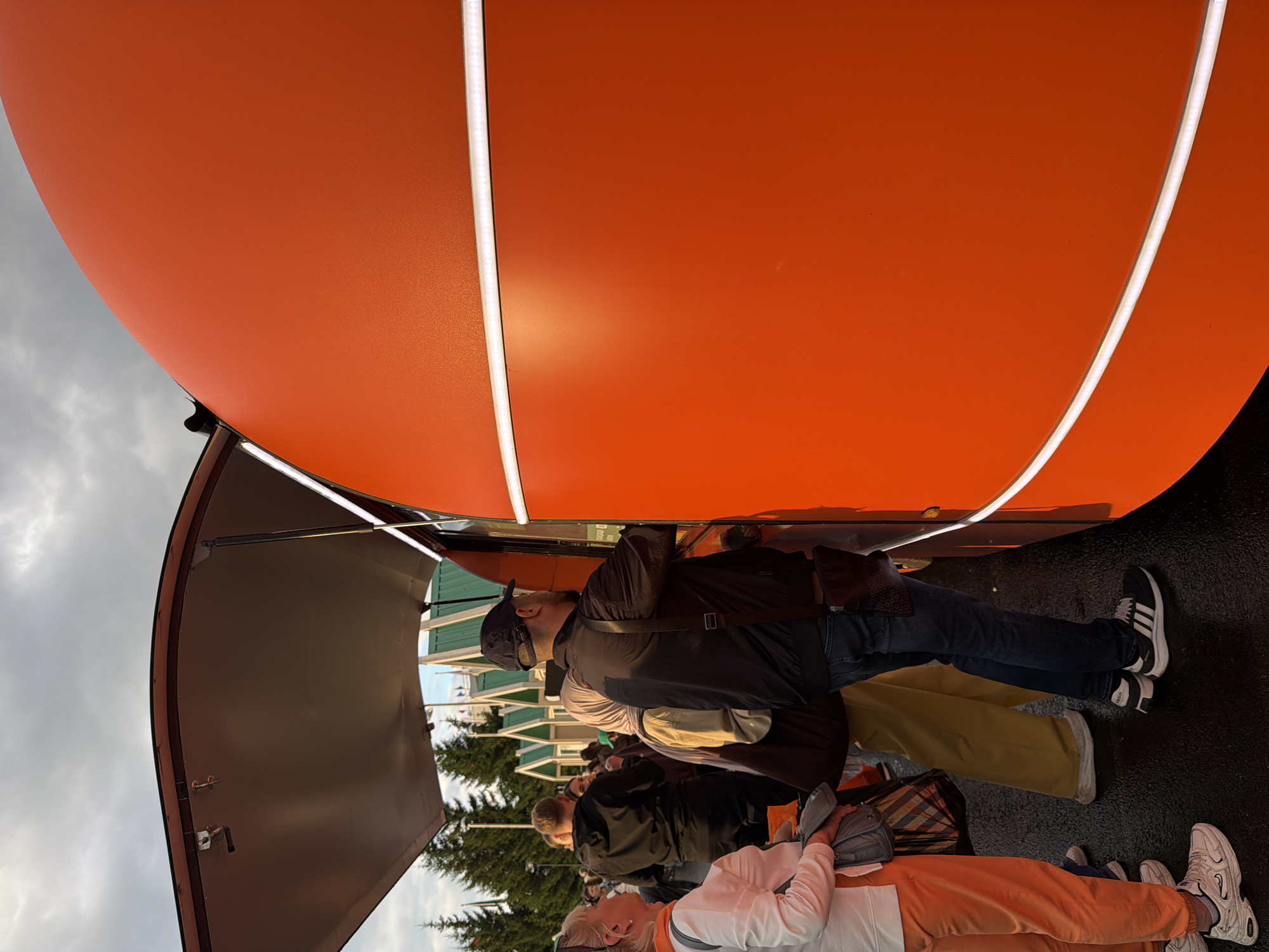The image depicts a group of people standing in front of a large, bright orange structure, possibly a food truck or a booth at an outdoor event. The structure has a curved top with prominent white LED strip lighting running along its edges. The scene appears to be outdoors, with a cloudy sky visible in the background. Trees can be seen behind the people, suggesting a park or outdoor venue.
The people are casually dressed, and their postures suggest they are waiting in line or interacting with the structure. One person in the foreground, facing away from the camera, is wearing a white jacket and orange pants, with sunglasses on their head. Another person in the middle ground, wearing a dark jacket and a baseball cap, is looking towards the orange structure. The lighting suggests it could be late afternoon or early evening, as the sky is overcast. The overall atmosphere seems relaxed and casual, typical of an outdoor gathering or market. There is no visible text in the image.