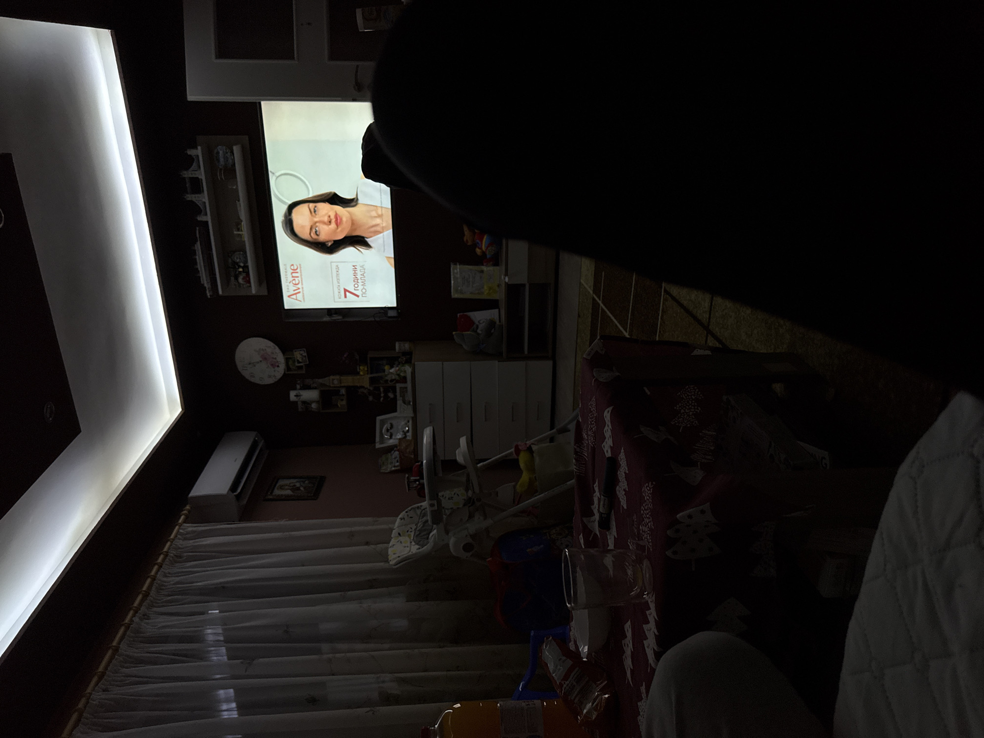 The image appears to be taken from an indoor setting, possibly a living room in Sofia, Bulgaria, considering the Bulgarian text visible on the television screen. A person is watching television, with an advertisement featuring a woman and the brand "Avène" displayed on the screen. The room seems dimly lit. In the foreground, there is a table covered with a Christmas-themed tablecloth, with various objects scattered on it including a drinking glass, bottles, and what appears to be a snack package. Parts of furniture, like a baby high chair, and other household items are vaguely visible in the background. The viewer's perspective seems to be from a reclined or relaxed position, potentially on a sofa or chair, with part of a blanket visible. The city of Sofia is inferred based on the Cyrillic text on the advertisement.