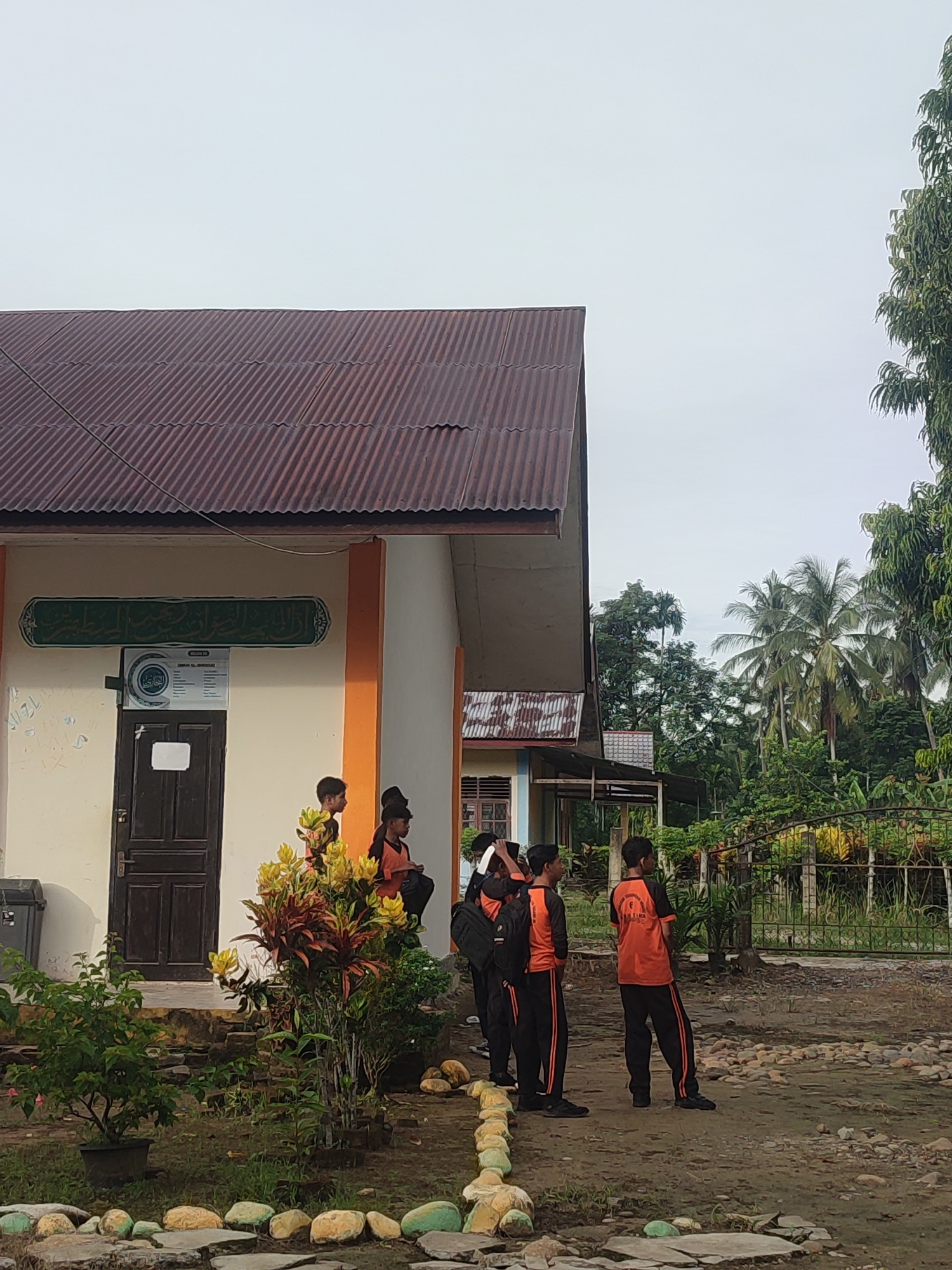 In Blangriek, Indonesia, a group of young males in school uniforms stand outdoors near a building. The building has a corrugated metal roof and a light-colored facade with an orange trim. A dark wooden door is visible on the facade, and a sign with Arabic script is mounted above it. One young man, wearing an orange t-shirt with black sleeves and black pants with an orange stripe, stands facing away from the camera. Another young man, similarly dressed, stands to his left. Other young men are visible in the background, some wearing similar uniforms, others in different attire. The ground is covered in dirt and scattered rocks. Lush green vegetation and palm trees are present in the background, suggesting a tropical environment. The sky is overcast and a light gray.