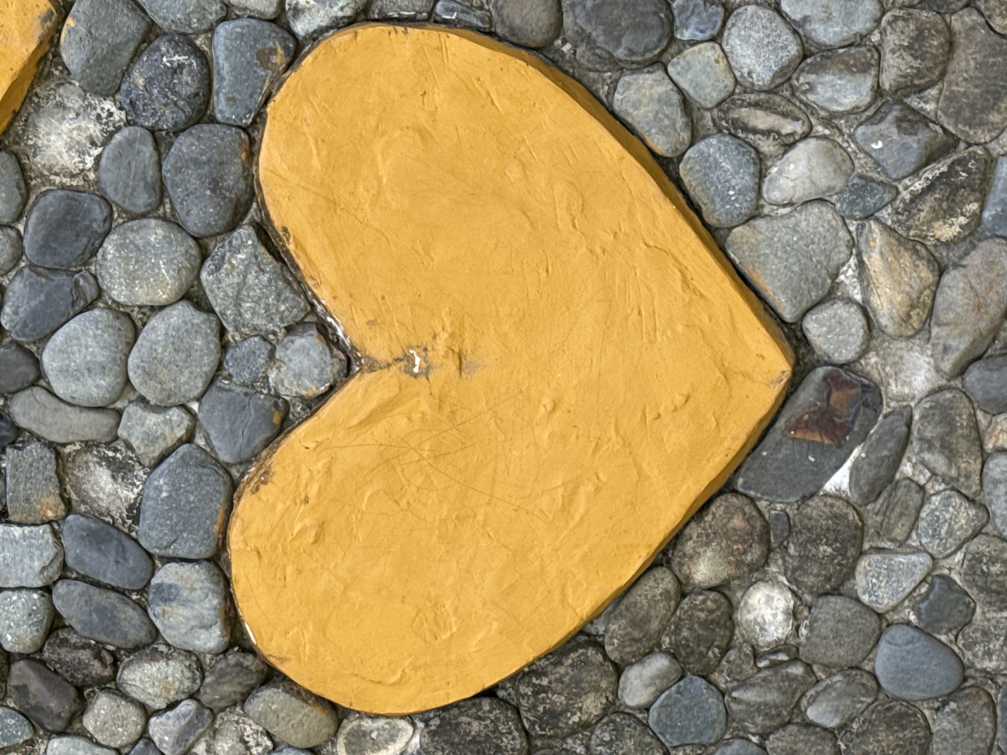 A close-up, overhead shot of a yellow, textured heart shape set into a wall of grey river stones. The heart is centrally located, taking up most of the frame. The stones are of varying sizes and shapes, with some smoother and rounder, while others are more angular and rough. The heart has a matte finish and appears to be made of painted concrete or stucco. The overall impression is one of rustic charm and simple decoration. The location is the city of Nam Khanh Vinh, Vietnam.