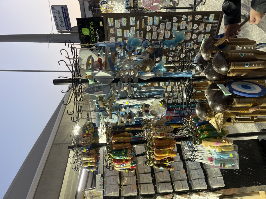 The image shows a souvenir stand in Limenaria, Greece (latitude: 40.626, longitude: 24.574). The stand is located on a street, with a building to the right and an awning overhead. It is filled with colorful souvenirs, including wooden fish, keychains, small wooden wind chimes, and small trinkets on display boards. A person is shown on the right side of the image, reaching toward the wind chimes. The stand is owned by "Greek Horizons Products." A sign for a restaurant "Restaurant - Grill Palataki" is visible on the building to the right.